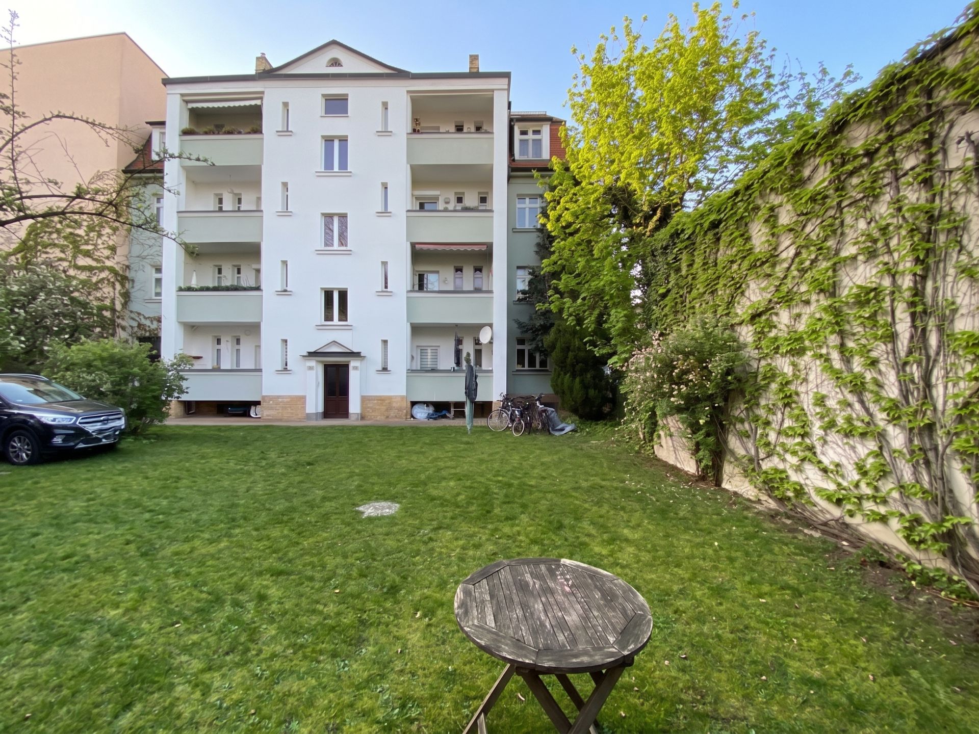 Wohngebäude mit mehreren Balkonen, davor Garten mit Rundtisch und Bäumen.