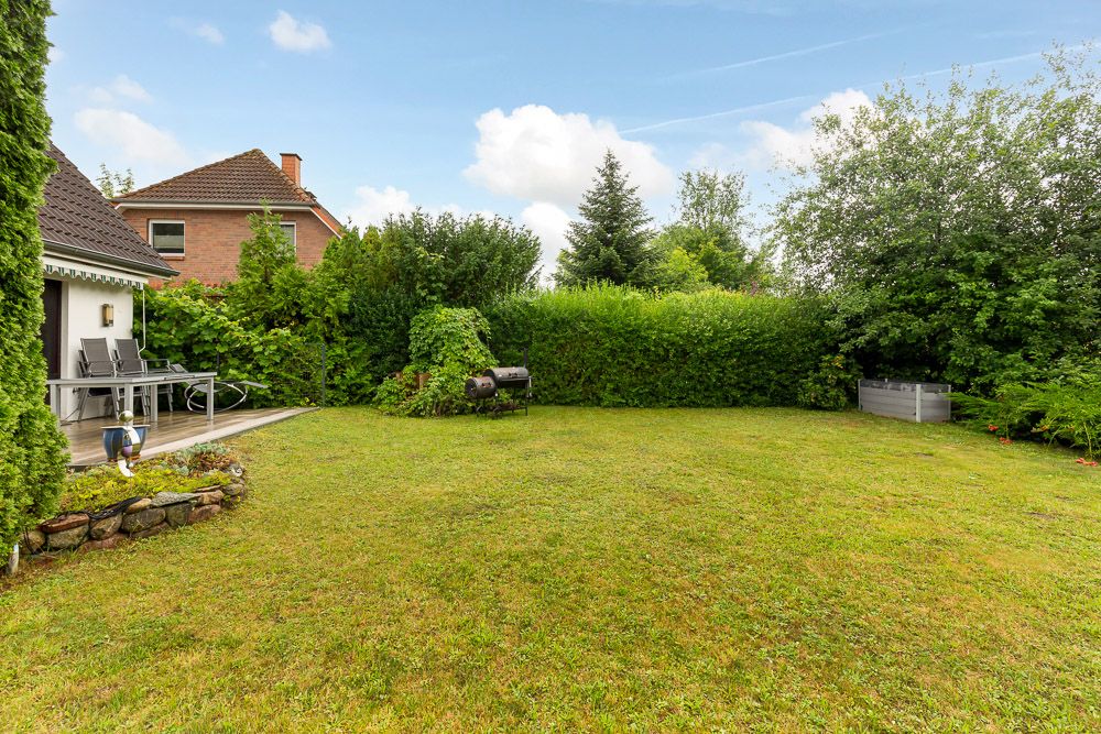 Grüner Garten mit Terrasse, Stühlen und Grill, umgeben von Hecken und Bäumen.