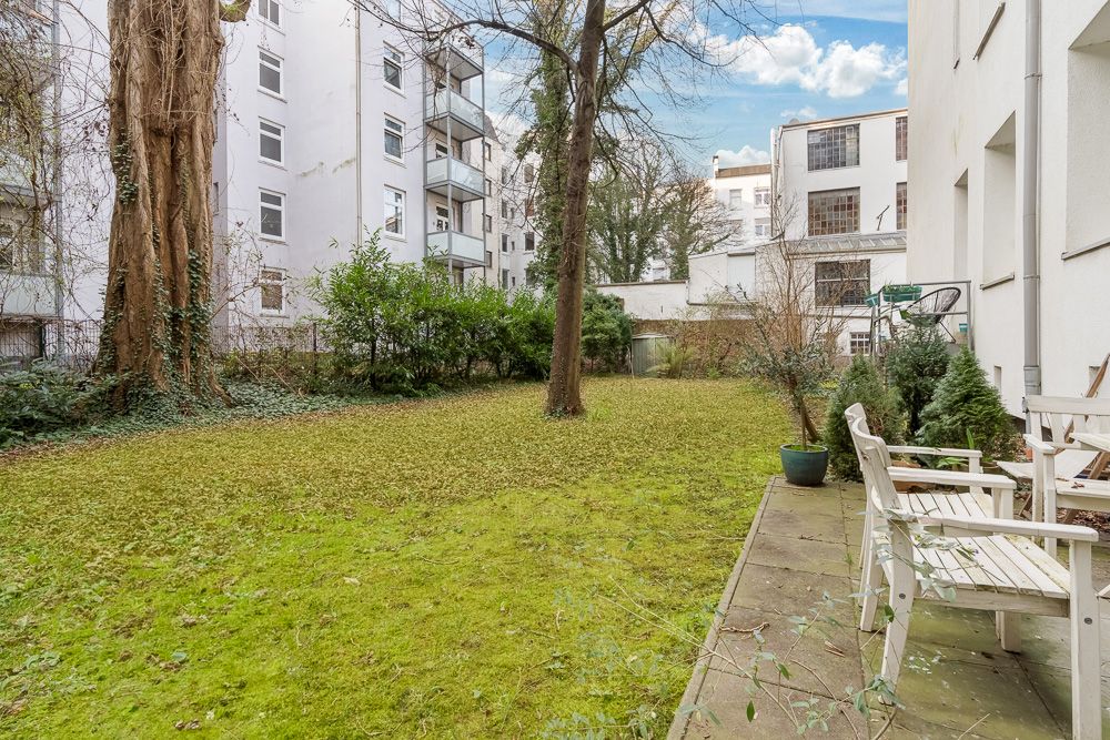 Grüner Garten mit Rasen, Baum, Sträuchern und Terrasse mit weißen Stühlen neben Wohngebäude.