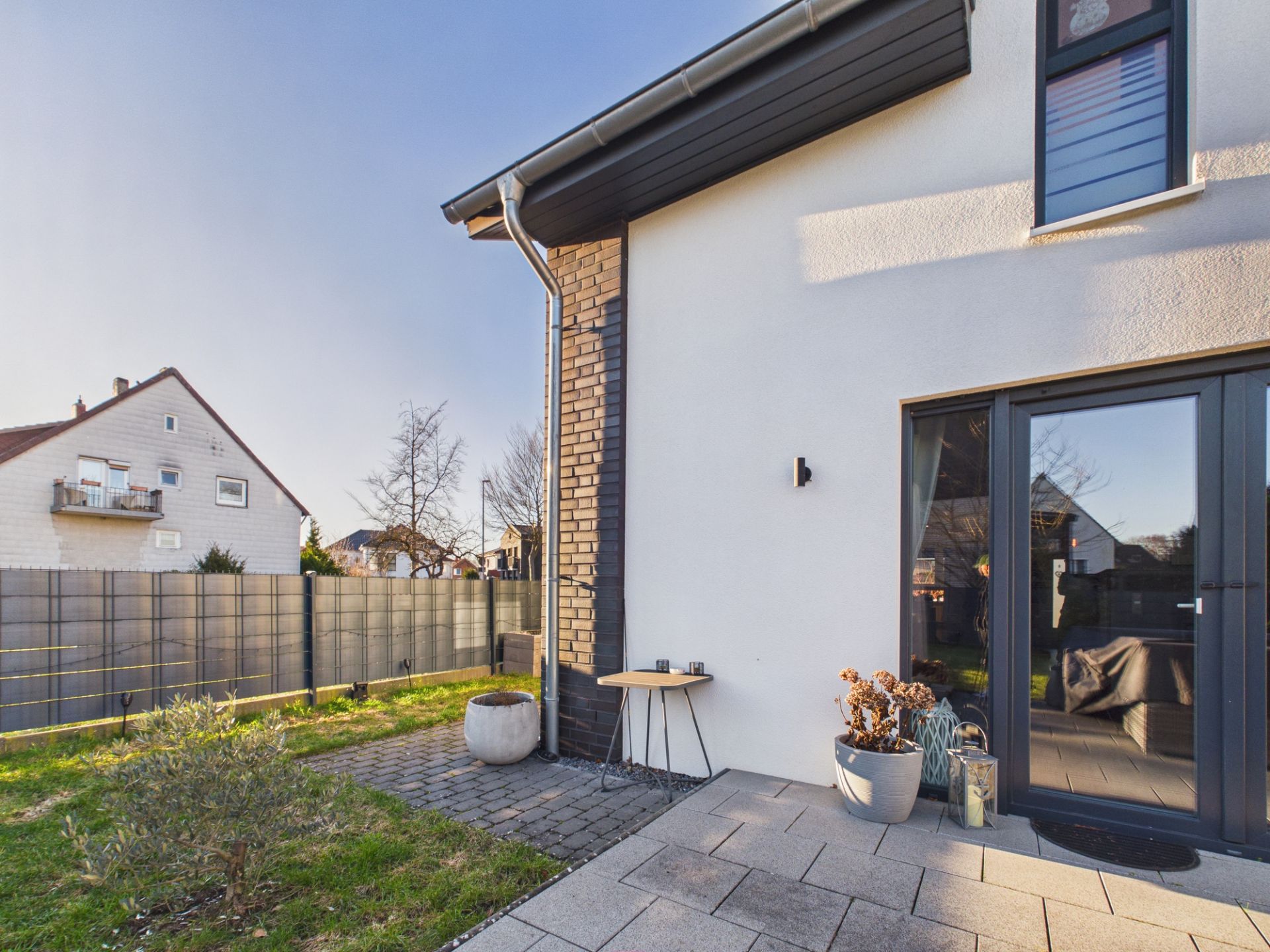 Moderne Hausfassade mit Glas-Schiebetür, Terrasse und zwei Blumentöpfen.