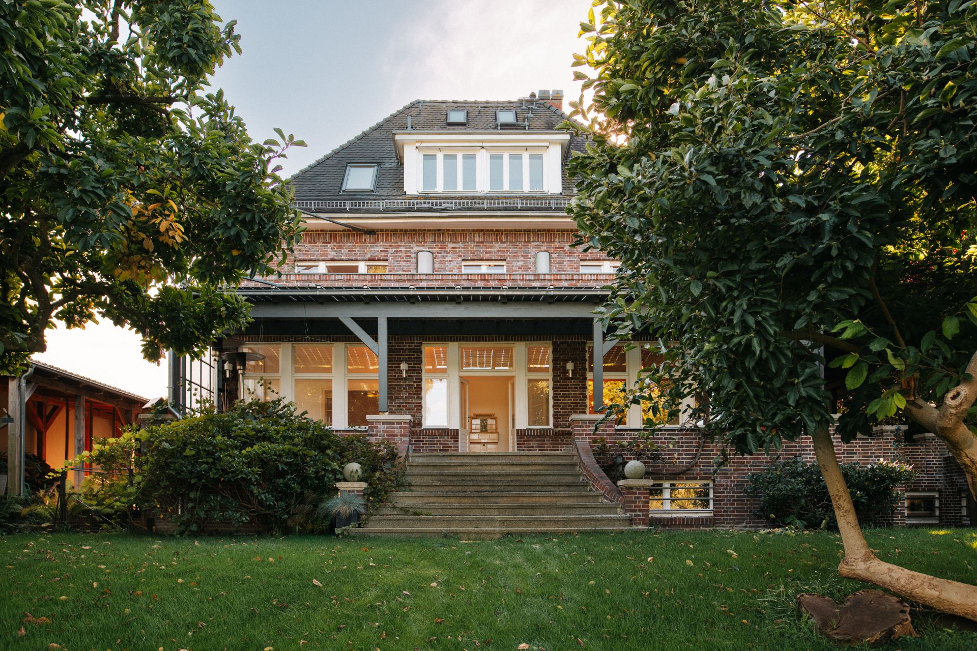 Backsteinwohnhaus mit großer Fensterfront und Treppe, umgeben von Bäumen und Garten.