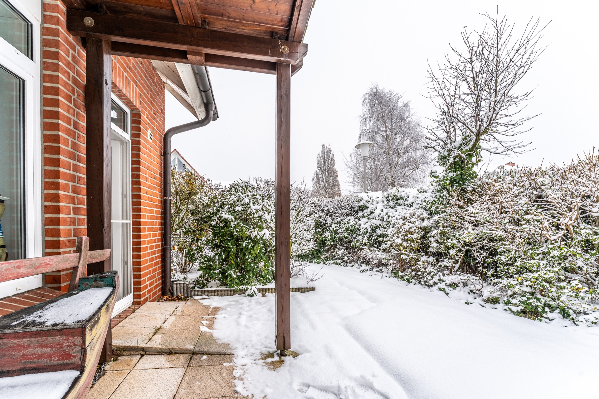 Verschneiter Garten mit Terrasse, Bank und Hecke neben einem Backsteinhaus.