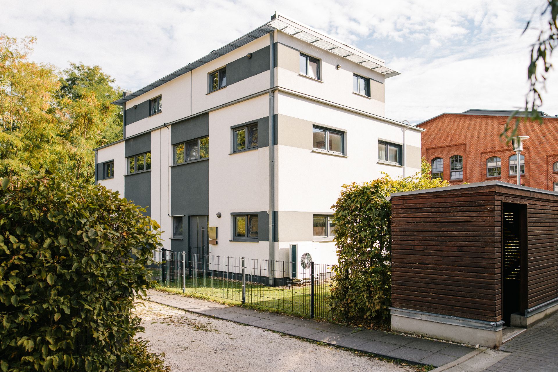 Modernes, dreistöckiges Wohnhaus mit flachem Dach und kleinem Garten, daneben ein Holzschuppen.