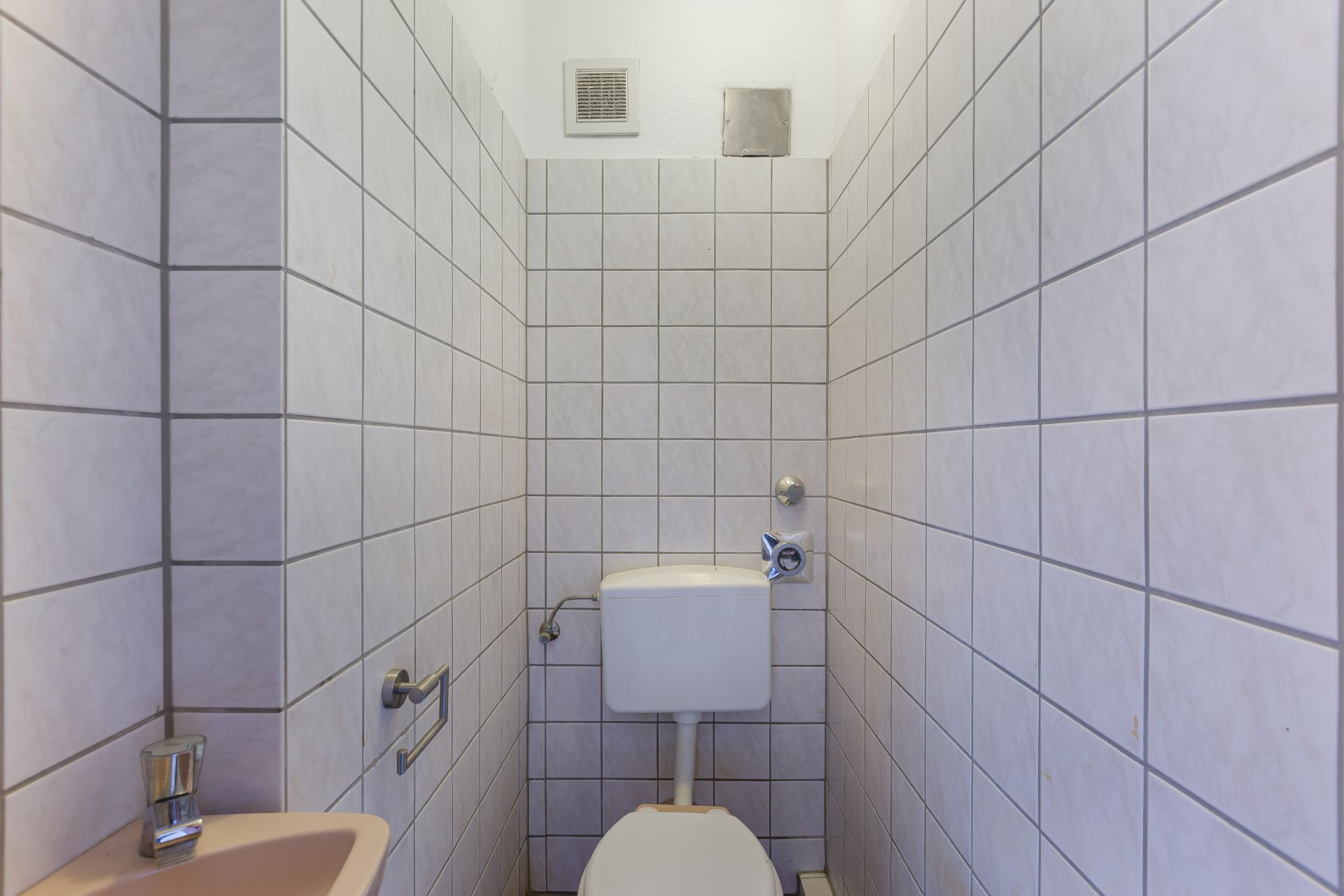 Schmaler gefliester Raum mit Toilette, Spülkasten und kleinem Waschbecken.