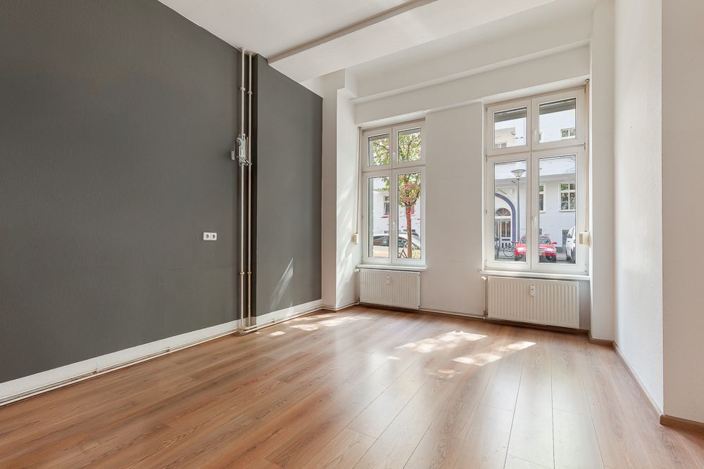 Helles, leeres Zimmer mit Holzfußboden, grauer Wand und zwei großen Fenstern.