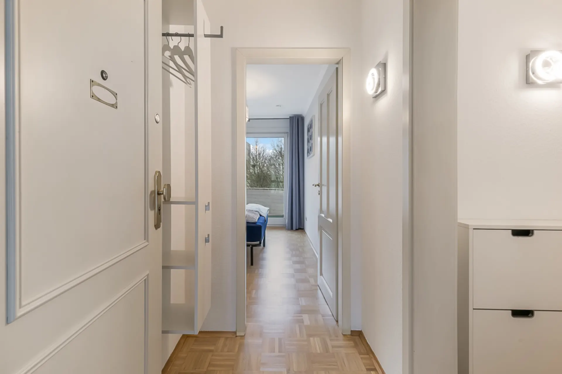 Heller Flur mit offener Tür, Kleiderhaken und Blick in ein Zimmer mit Fenster.