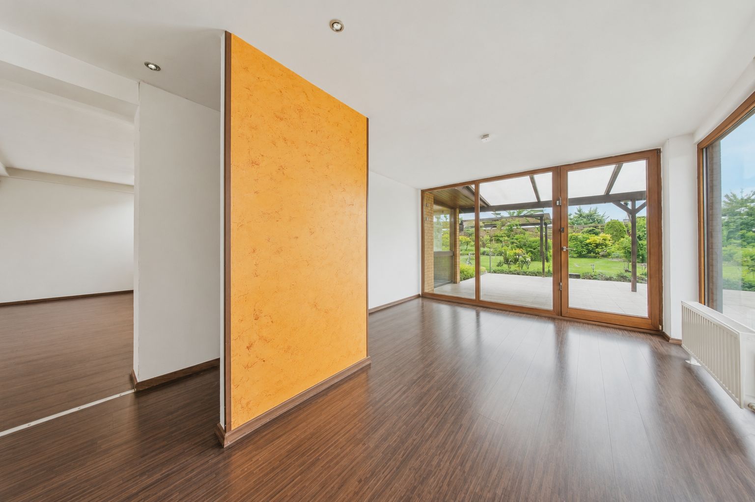 Helles Zimmer mit Holzfußboden, gelber Trennwand und großen Fenstern mit Blick in den Garten.