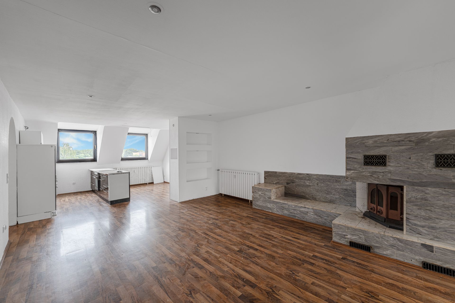 Heller Raum mit Holzboden, zwei Fenstern, offener Küche und gemauertem Kamin.
