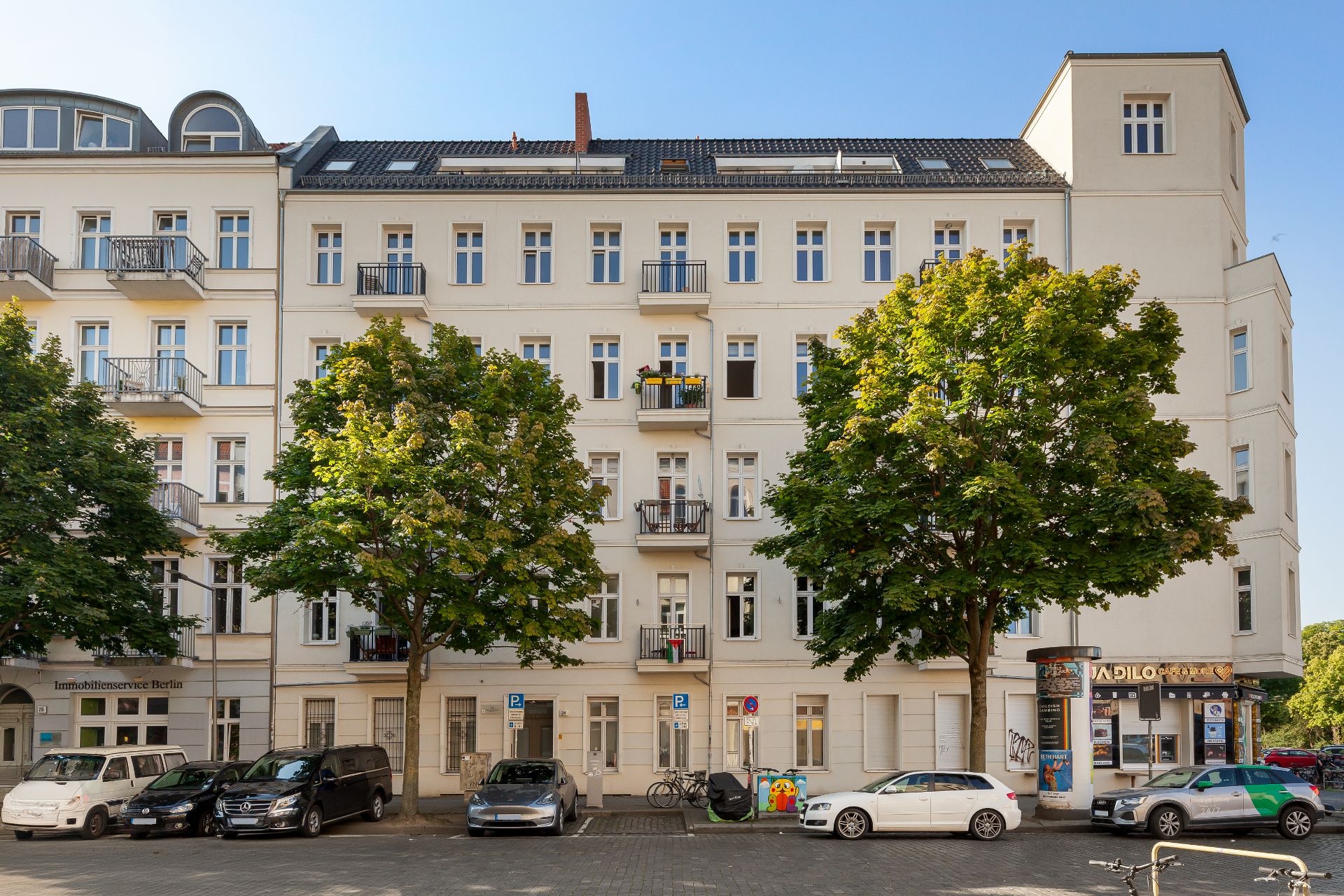 Mehrstöckiges Wohnhaus mit Balkonen, Bäumen und parkenden Autos davor.