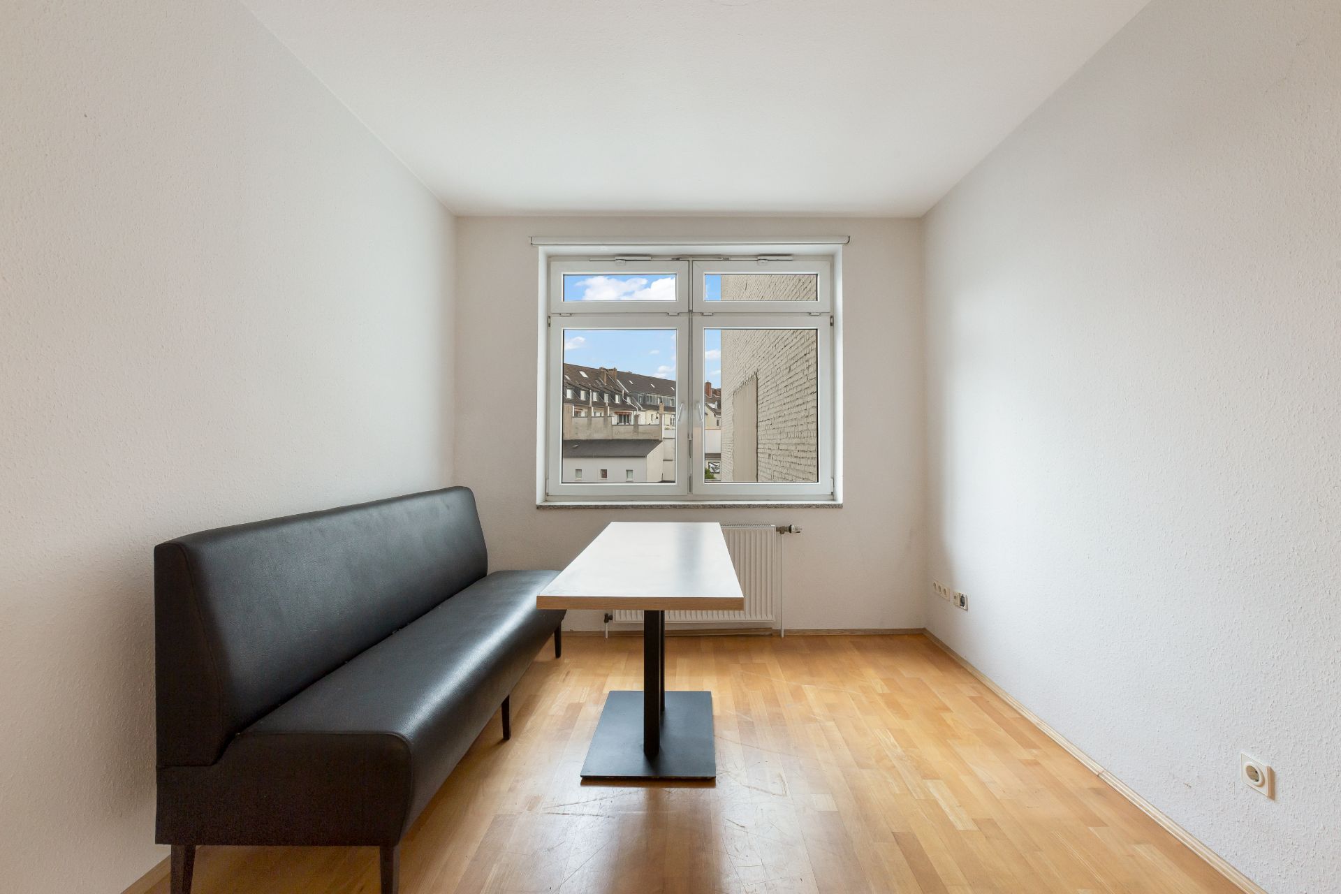 Helles Zimmer mit Holzboden, schwarzer Bank, Tisch und Fenster mit Blick nach draußen.
