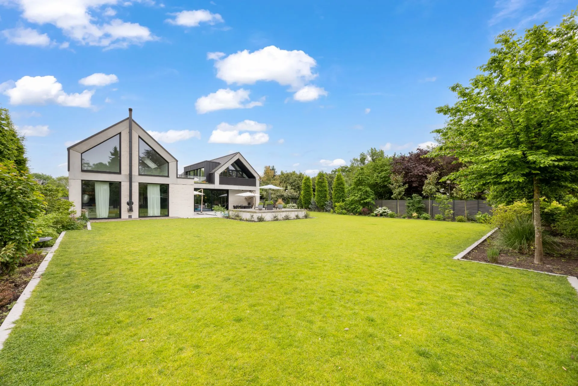 Modernes Haus mit großen Fenstern und weitläufigem Garten, umgeben von Bäumen und Sträuchern.