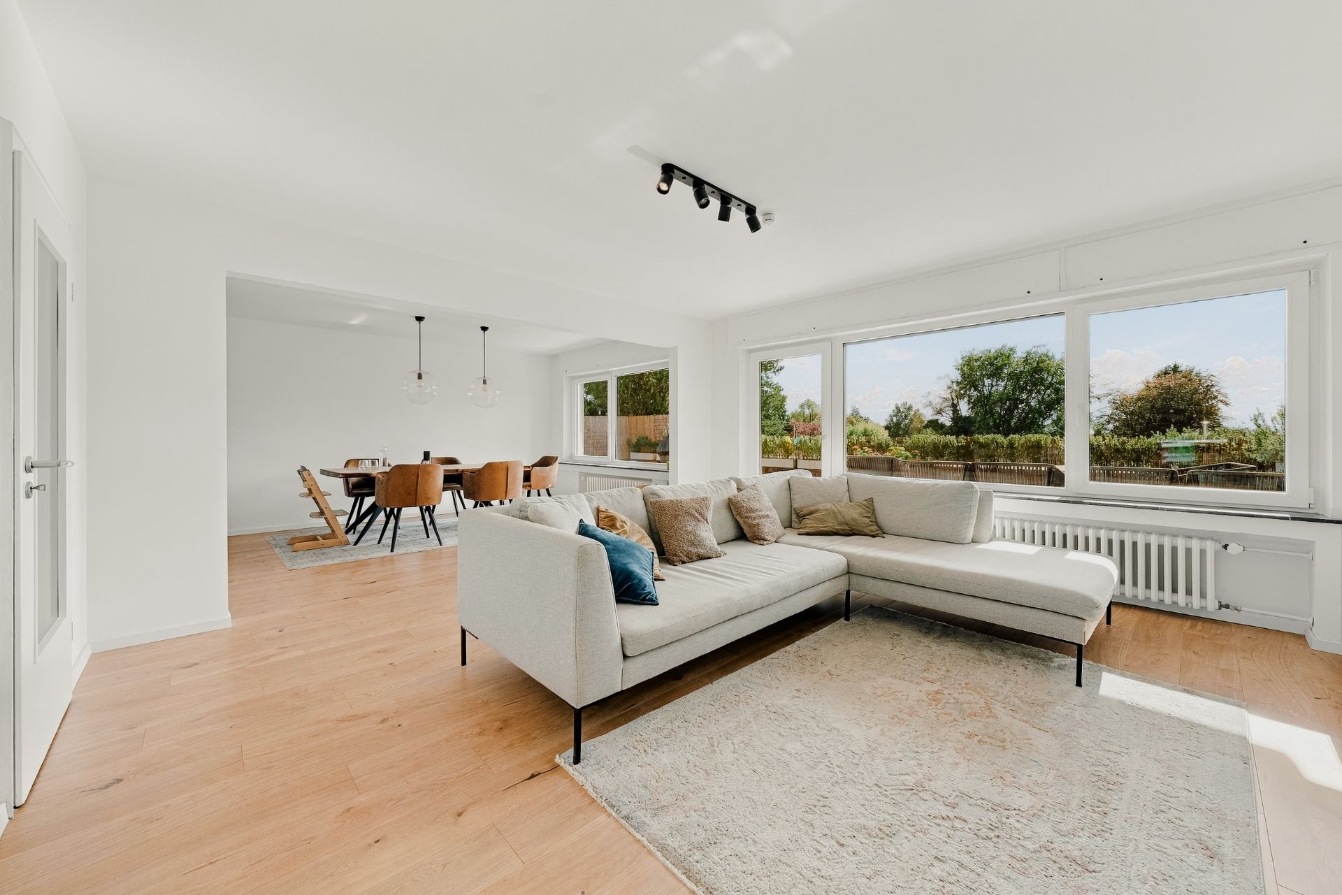 Helles Wohnzimmer mit Ecksofa, großem Teppich und Essbereich mit Tisch und Stühlen, große Fenster mit Blick ins Grüne.