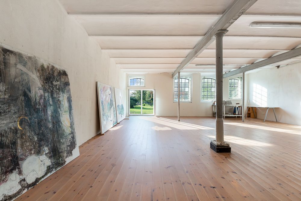 Großer, heller Raum mit Holzboden, großen Fenstern, Malereien an der Wand und Tischen im Hintergrund.