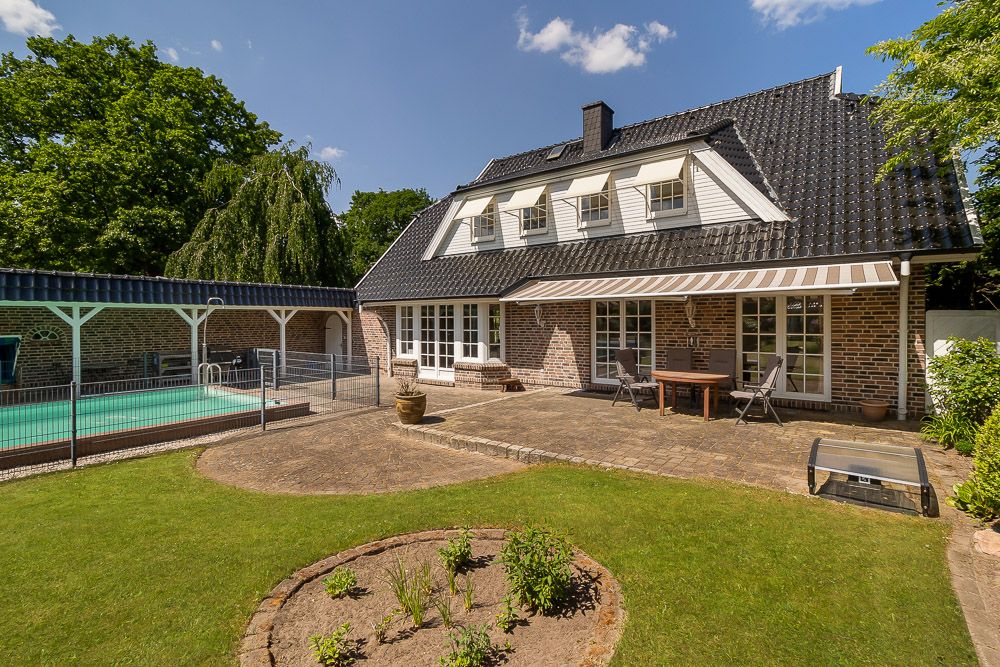Gepflegter Garten mit Terrasse, Tisch und Stühlen, daneben ein umzäunter Pool und ein Ziegelhaus.