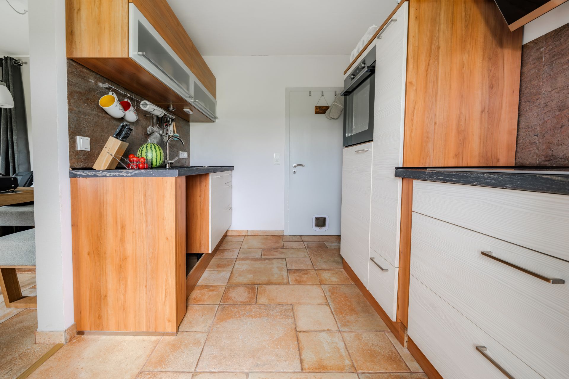 Moderne Küche mit Holzmöbeln, Hängeschränken und gefliestem Boden, Blick auf eine weiße Tür.