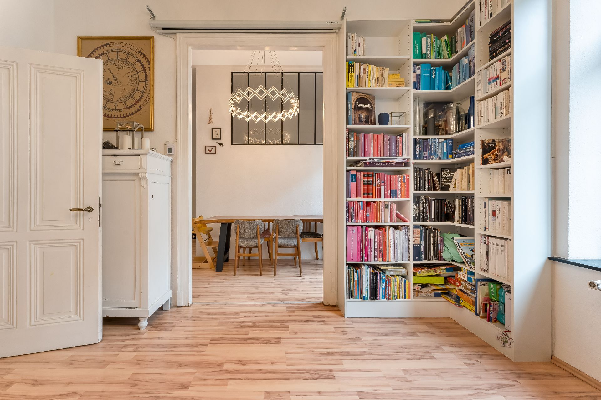 Helles Wohnzimmer mit Bücherregal, Holzboden und offenem Blick zum Essbereich mit Tisch und Stühlen.