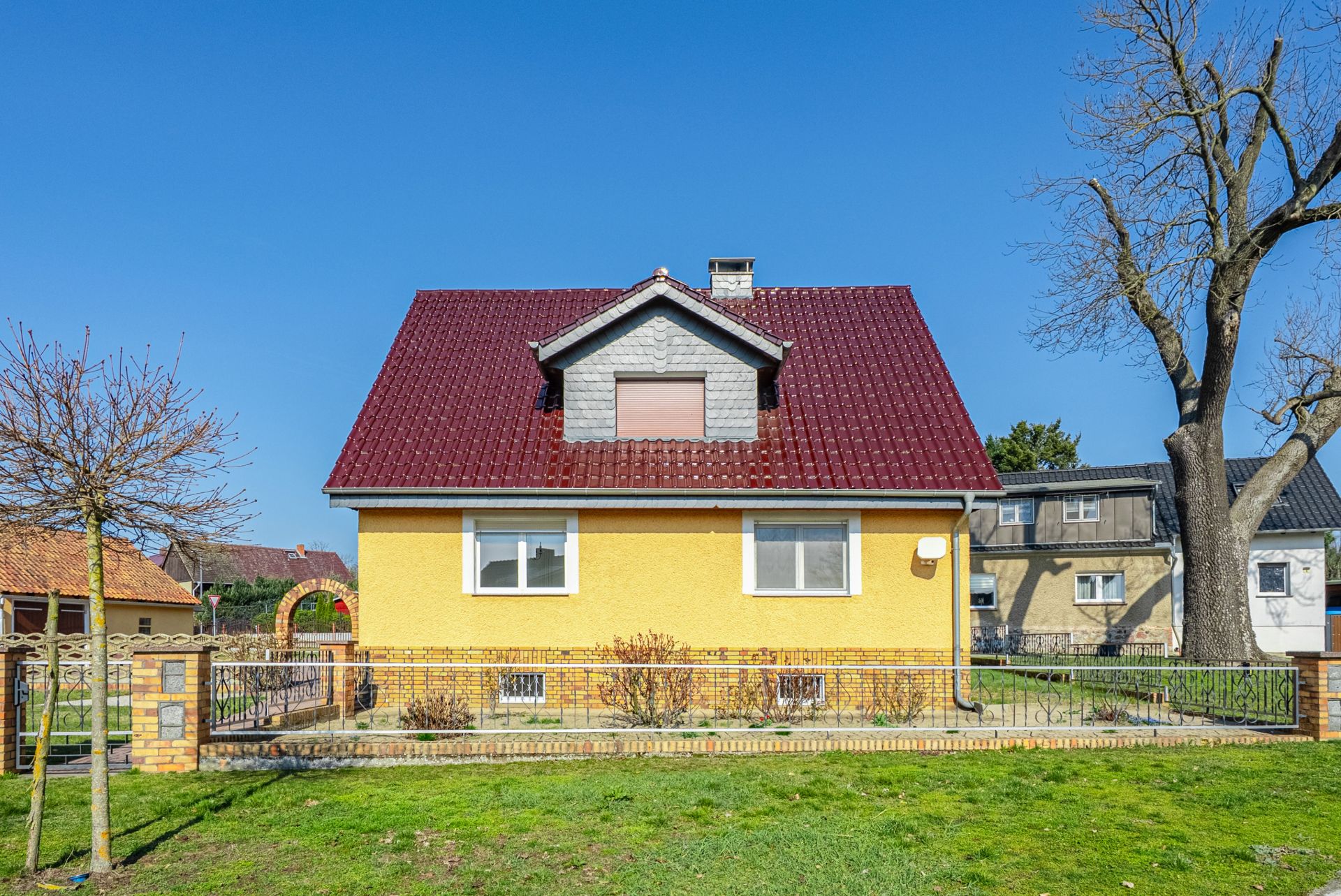 Gelbes Einfamilienhaus mit rotem Dach, Vorgarten und Zaun, Bäume an den Seiten.