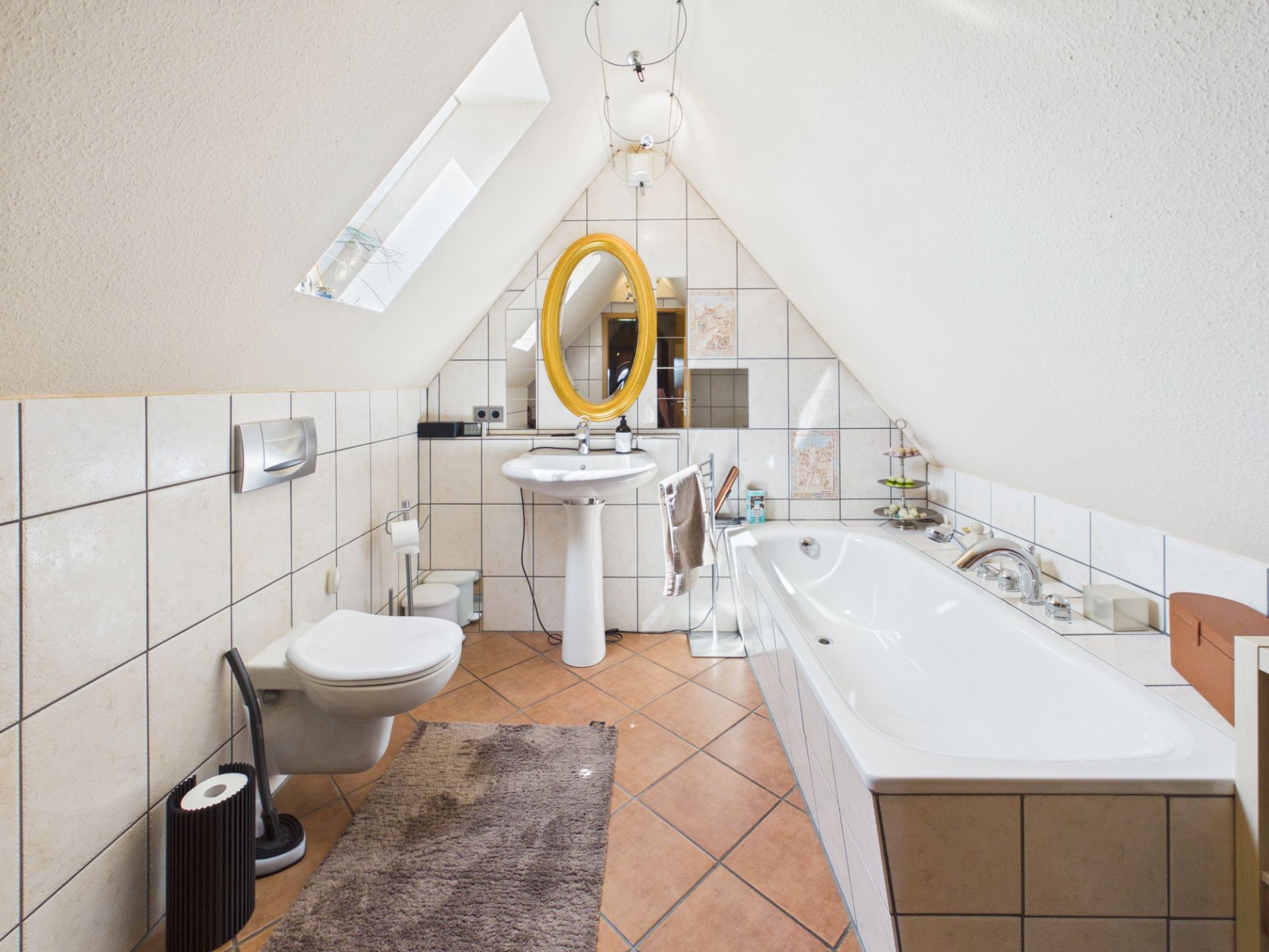 Helles Bad unter dem Dach mit Badewanne, Toilette, Waschbecken und rundem Spiegel.