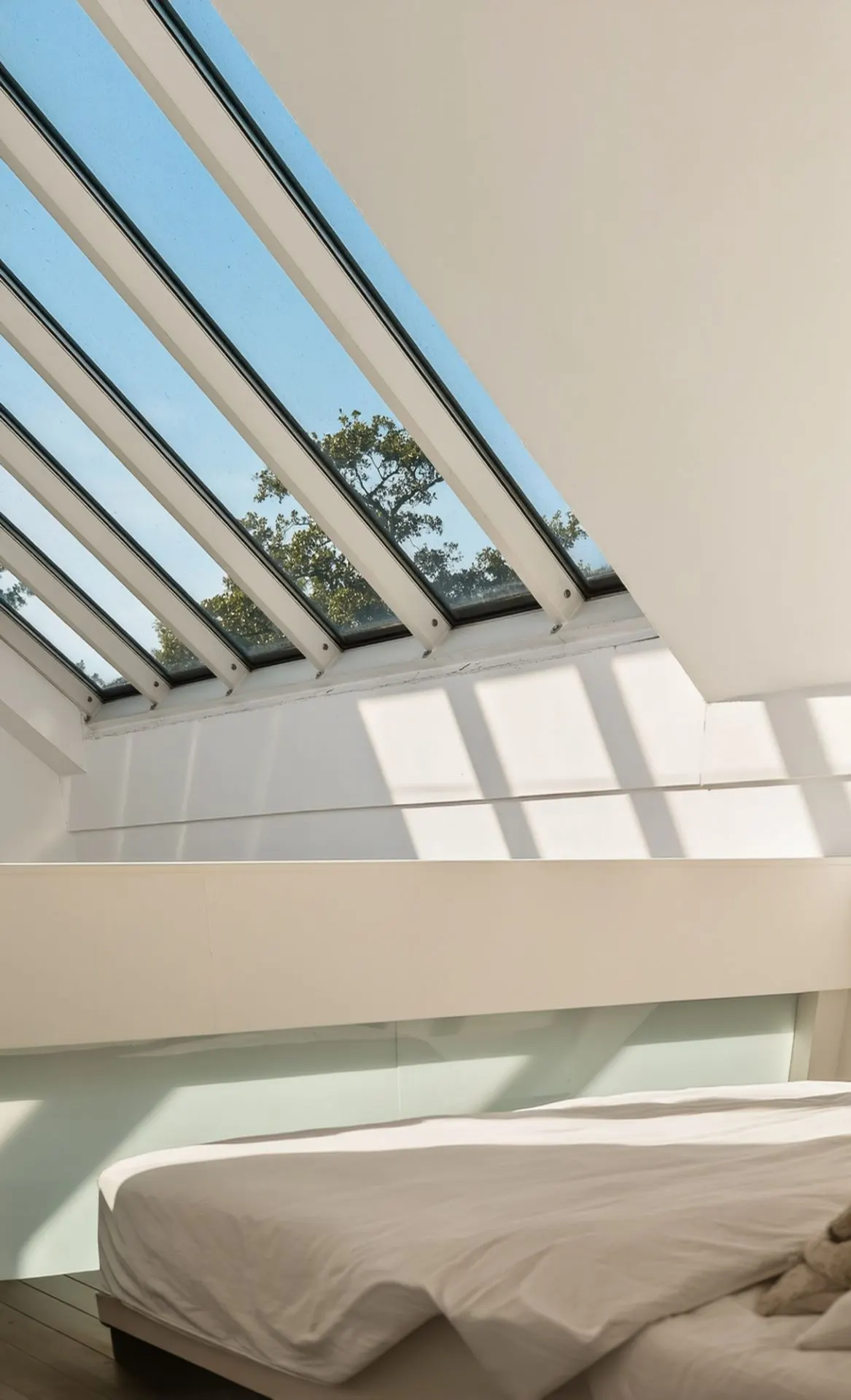 Modernes Schlafzimmer mit großem Schrägfenster und Blick auf Bäume, Sonnenlicht fällt auf das Bett.