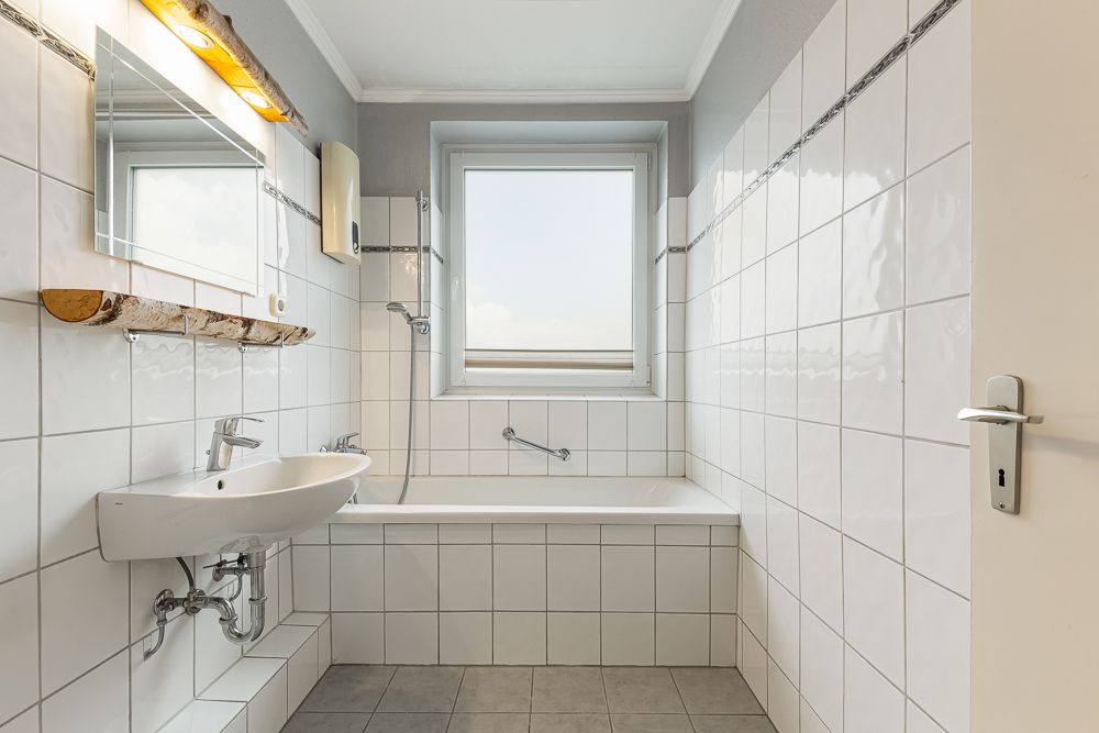 Helles Badezimmer mit Badewanne, Waschbecken, Spiegel und Fenster im Hintergrund.