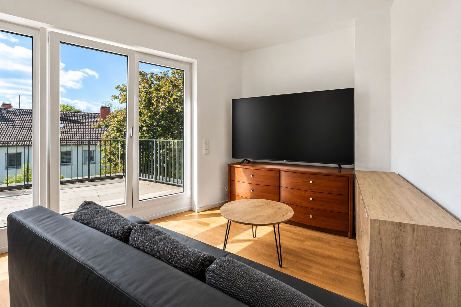 Helles Wohnzimmer mit Sofa, rundem Tisch, Fernseher und Zugang zum Balkon.