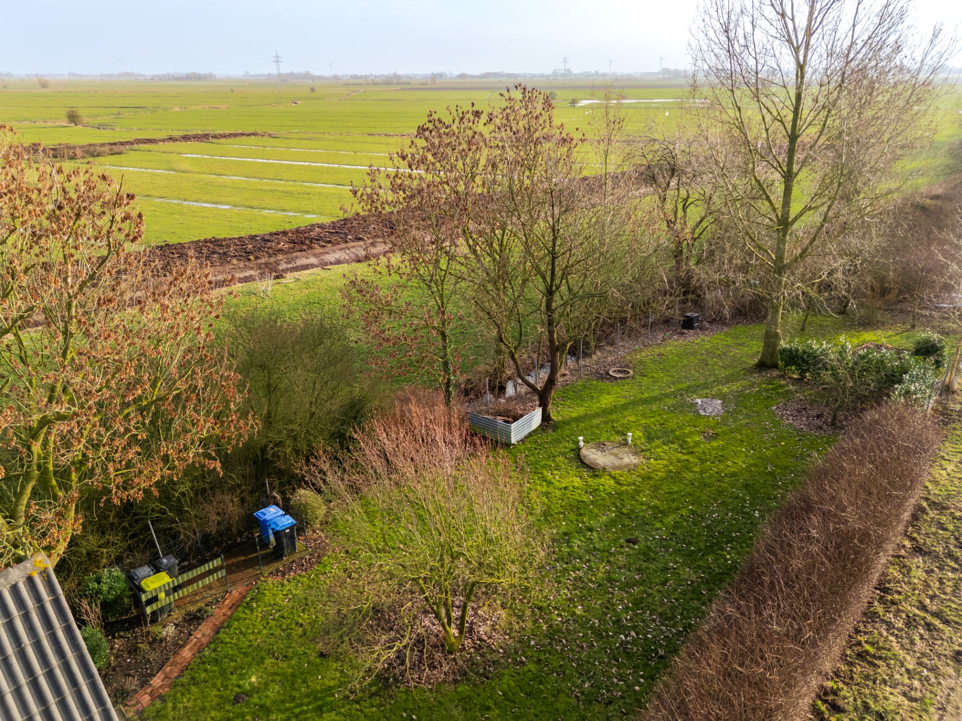 Grüner Garten mit Bäumen, Rasen und einzelnen Beeten, angrenzend an weite Felder.