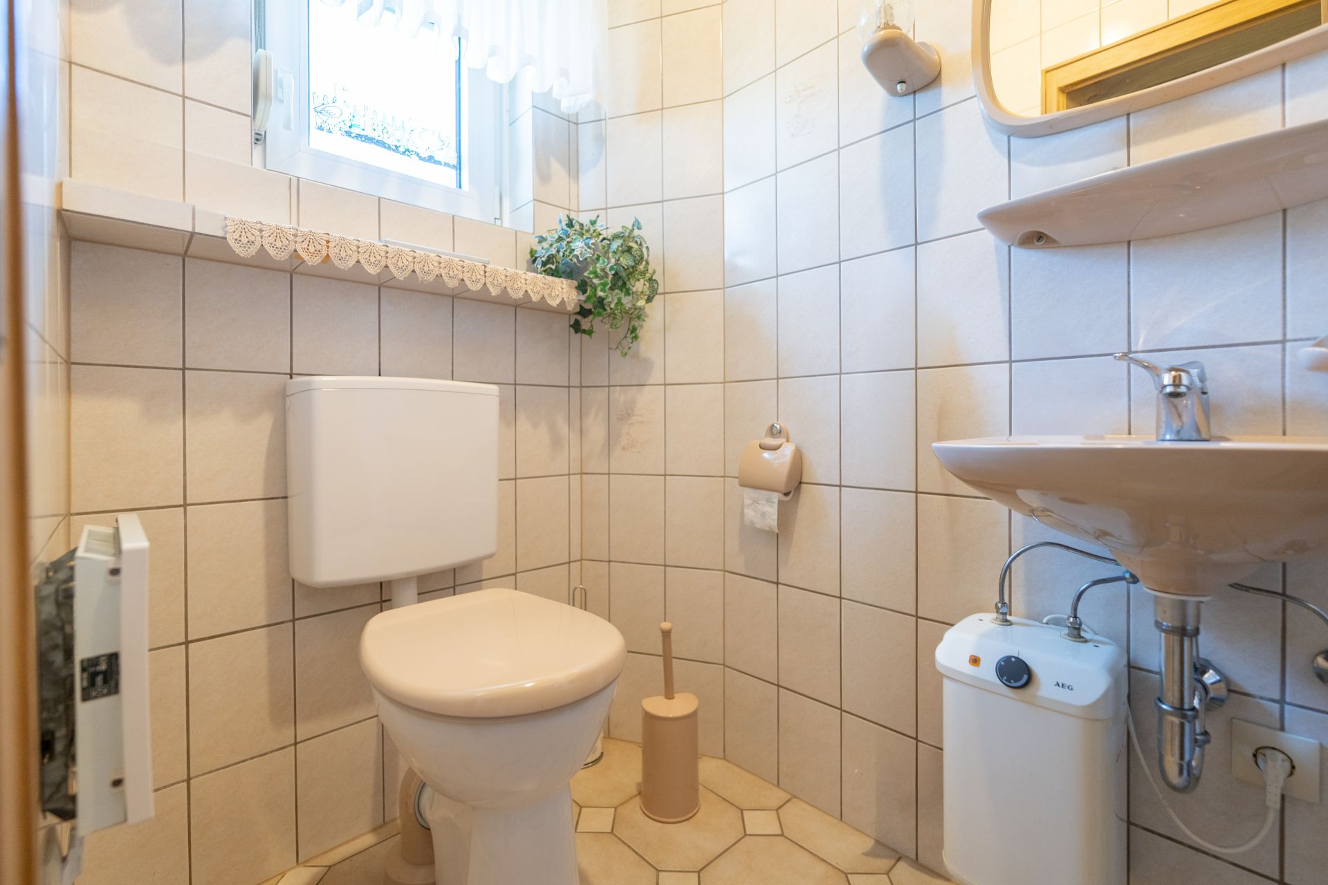 Kleines Badezimmer mit Toilette, Waschbecken, Spiegel, Fenster und einer Pflanze auf der Fensterbank.