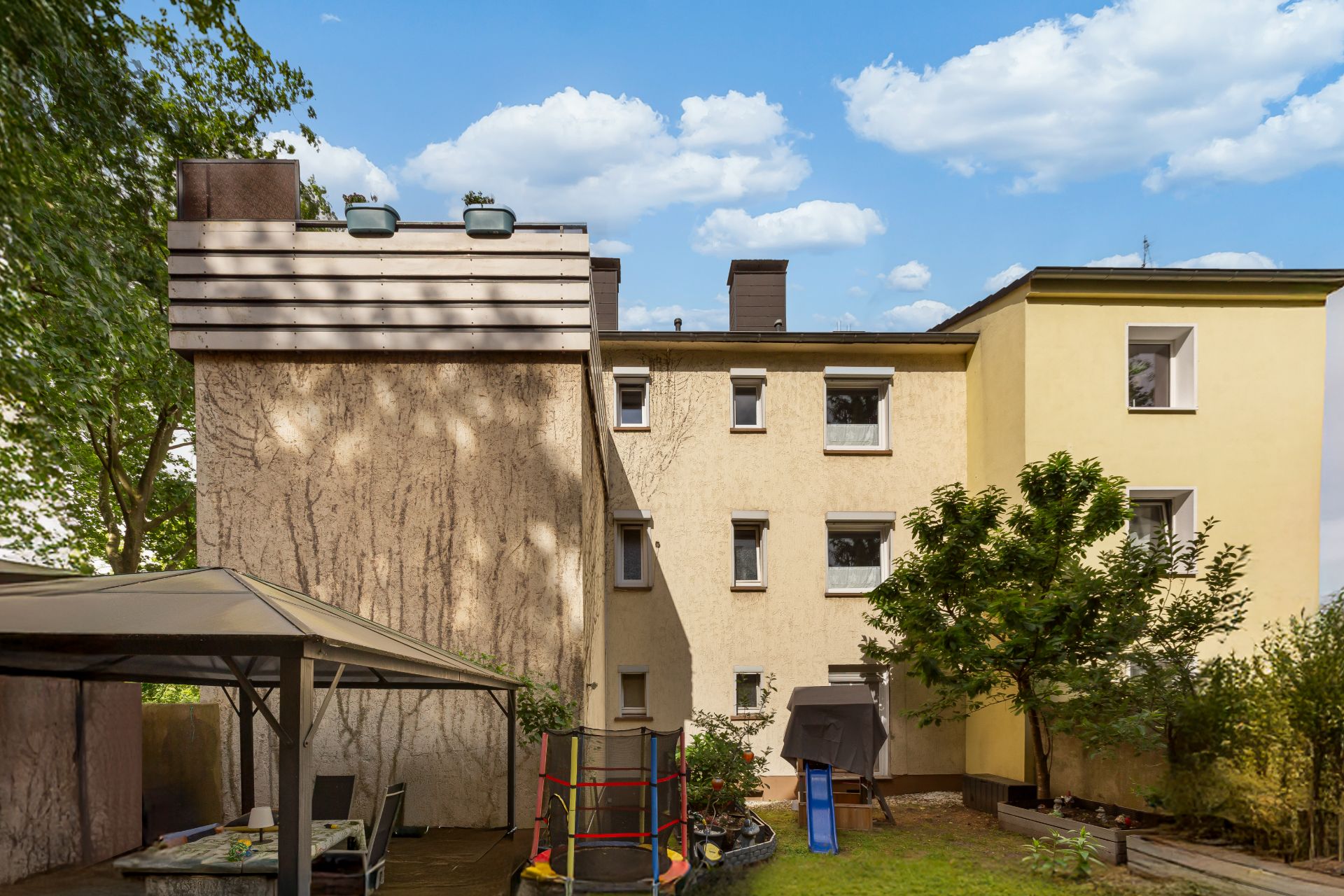 Hinterhof eines Wohnhauses mit überdachter Sitzfläche, Trampolin und Bäumen.