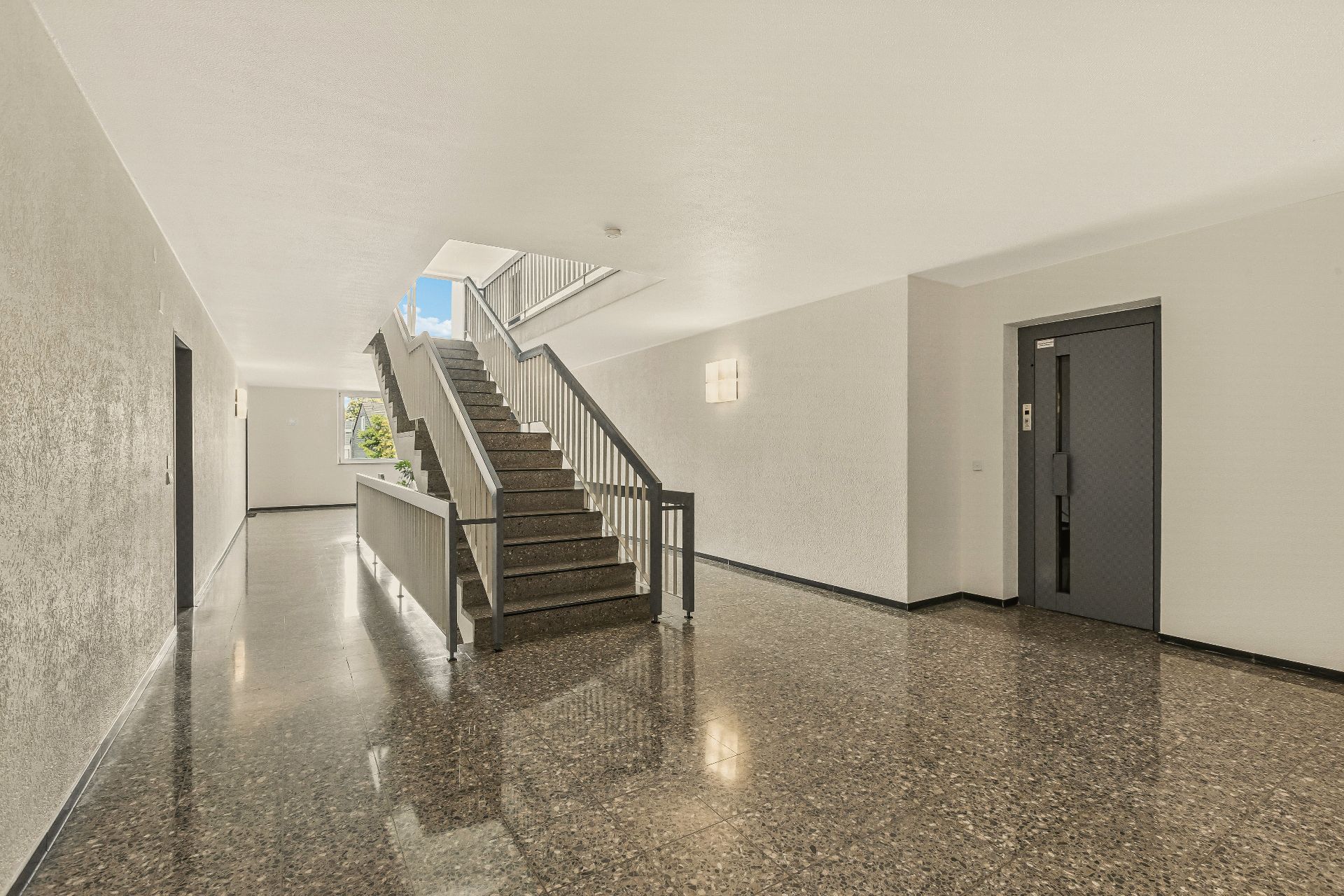 Heller Flur mit Treppenhaus, Aufzug und Fenster am Ende des Gangs.