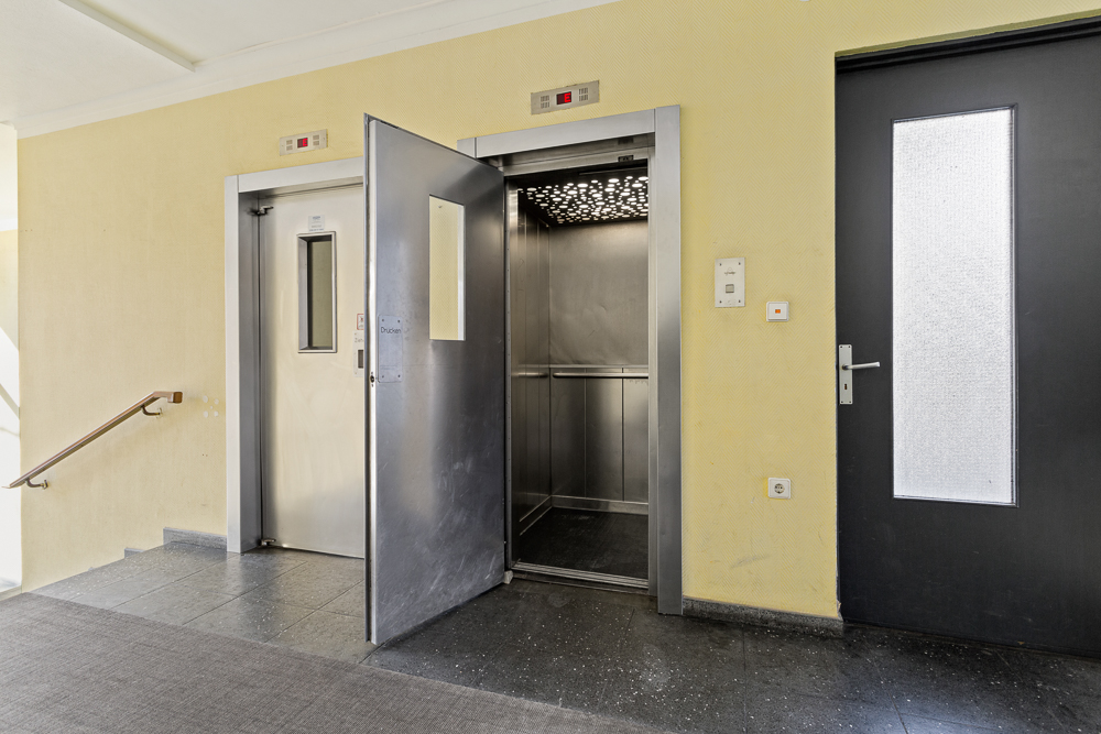 Offener Aufzug mit Edelstahltüren in einem Flur mit gelber Wand und schwarzer Tür.