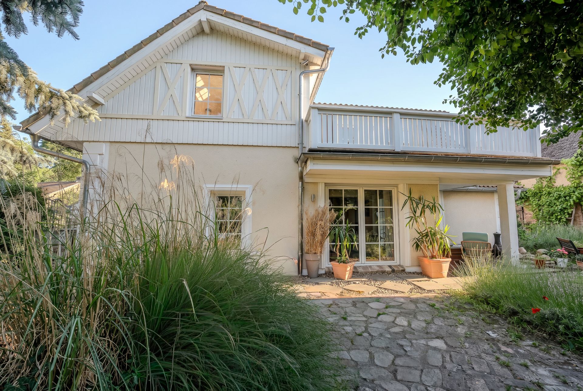 Zweistöckiges Haus mit Balkon, großer Fensterfront und Pflanzen im Garten.