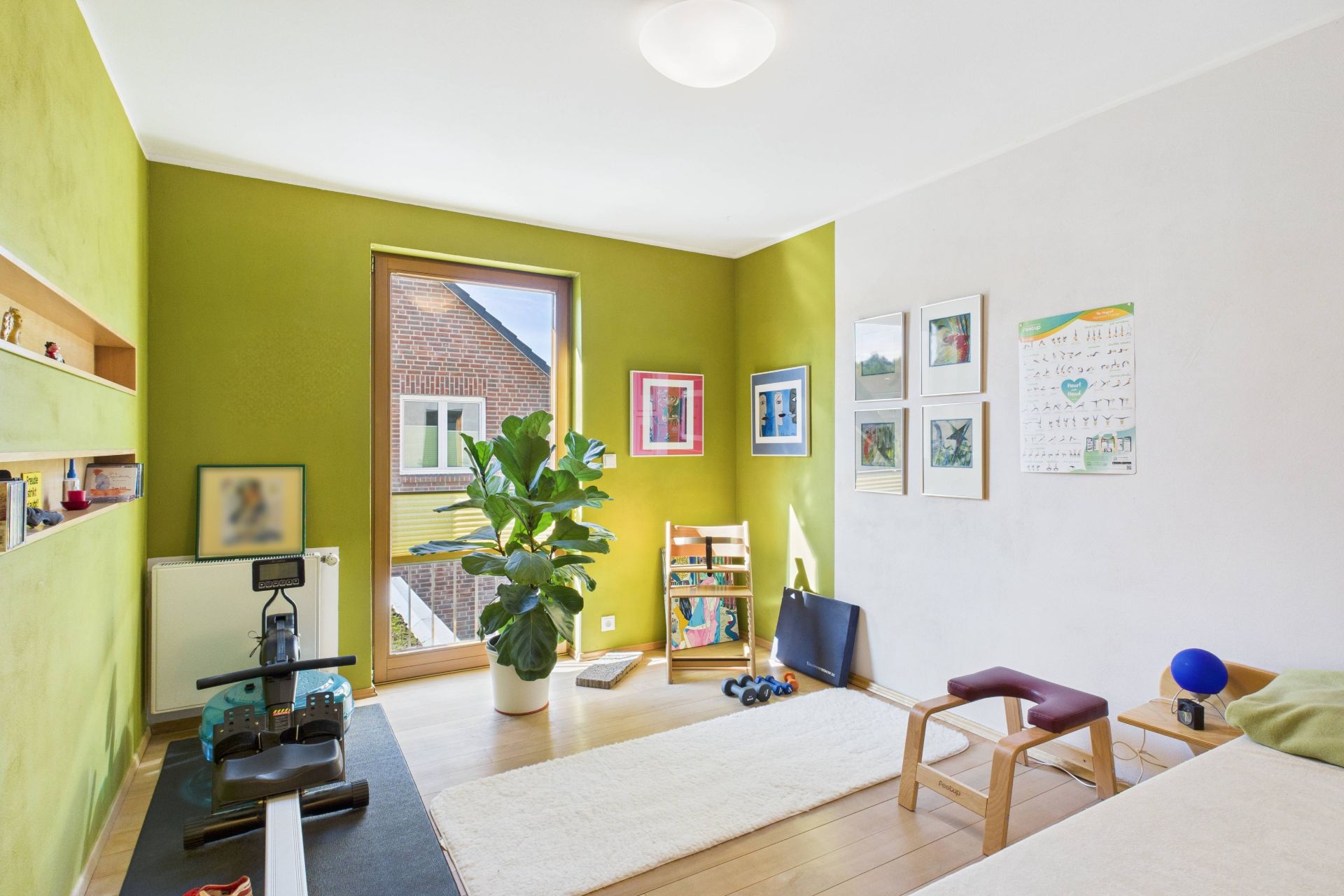 Helles Zimmer mit grün gestrichener Wand, Pflanzen, Fitnessgeräten und Bildern an der Wand.