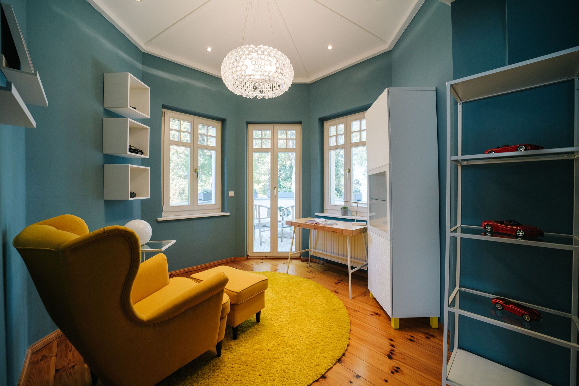 Helles Zimmer mit gelbem Sessel, Teppich, Schreibtisch, Regal und großen Fenstern.