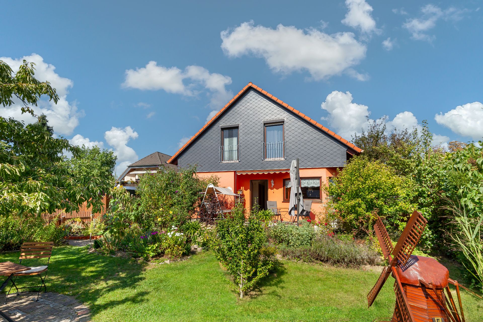 Einfamilienhaus mit Garten, Gartenmöbeln und kleiner Holzmühle bei sonnigem Wetter.