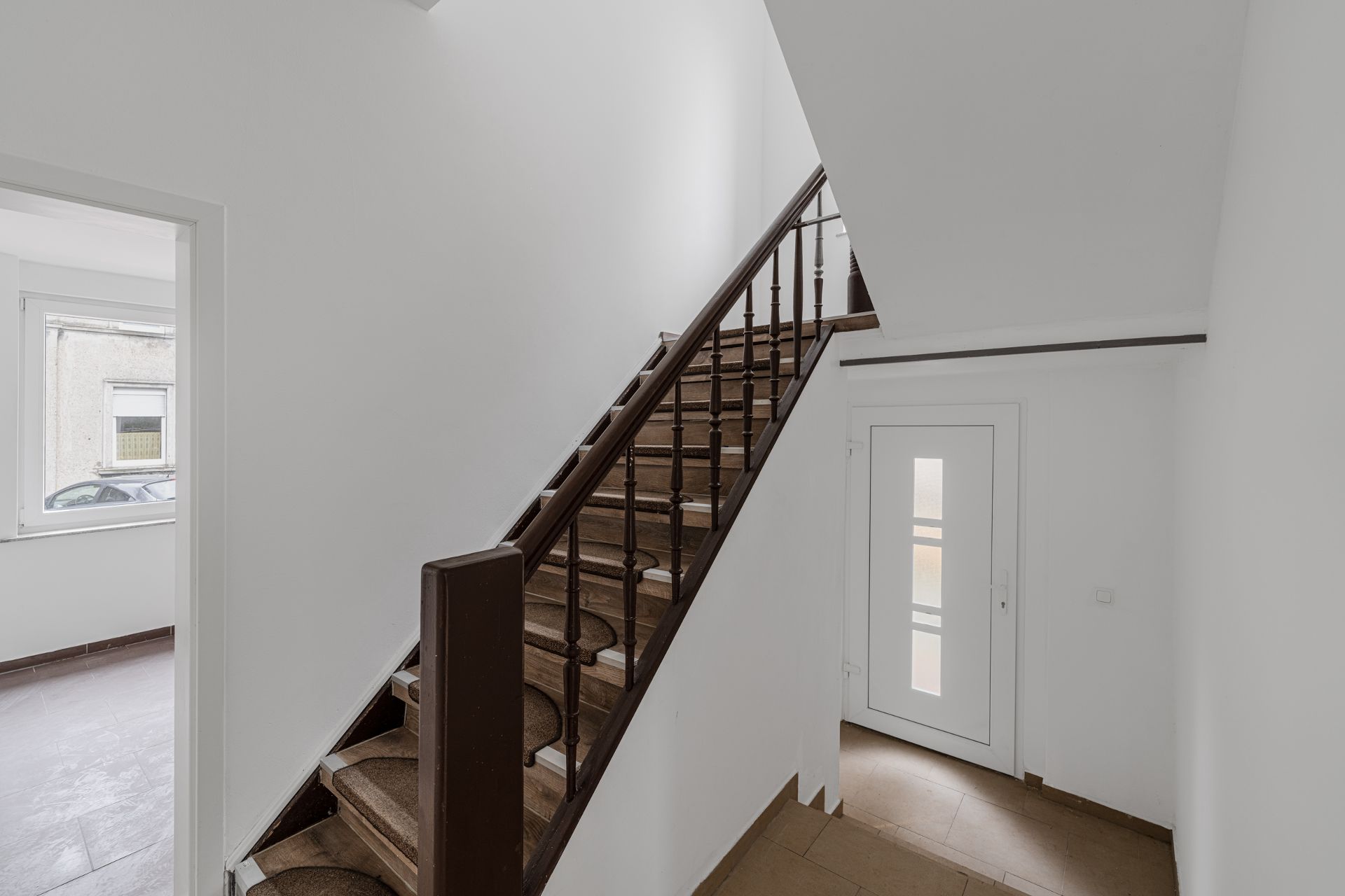 Heller Flur mit Treppe, weißer Tür und Zugang zu einem Raum mit Fenster.