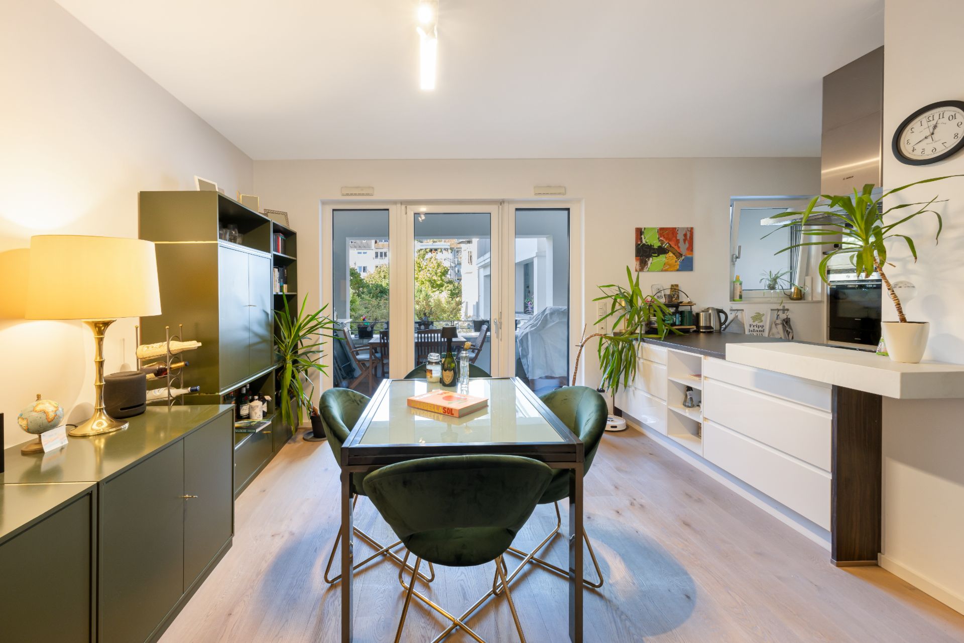Heller Essbereich mit Glastisch, grünen Stühlen, Pflanzen und Zugang zu einem Balkon.