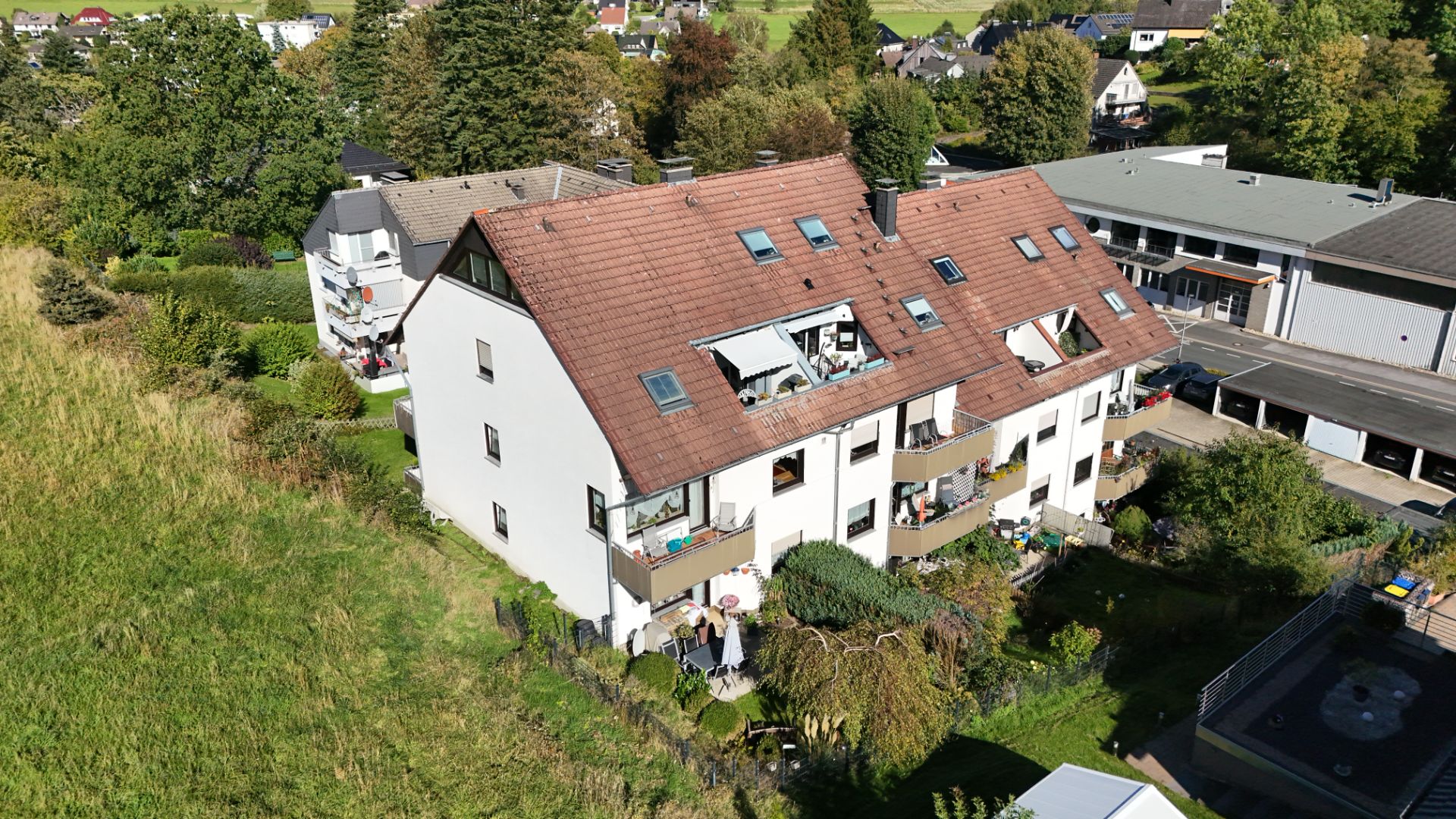Mehrfamilienhaus mit braunem Satteldach, Balkonen und umgebenden Gärten bei sonnigem Wetter.