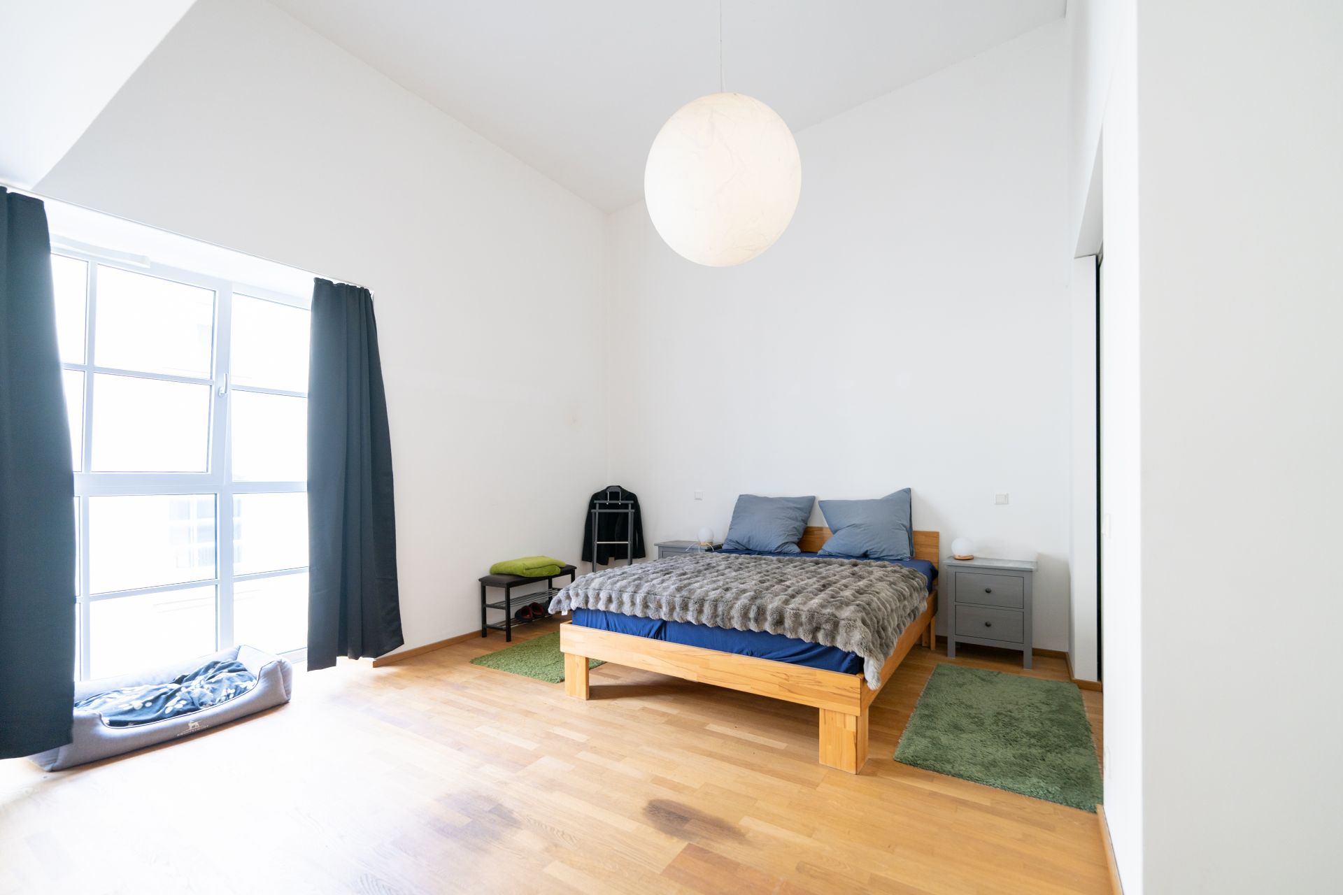 Helles Schlafzimmer mit Holzboden, Doppelbett, Nachttisch, Fenster und dunklen Vorhängen.