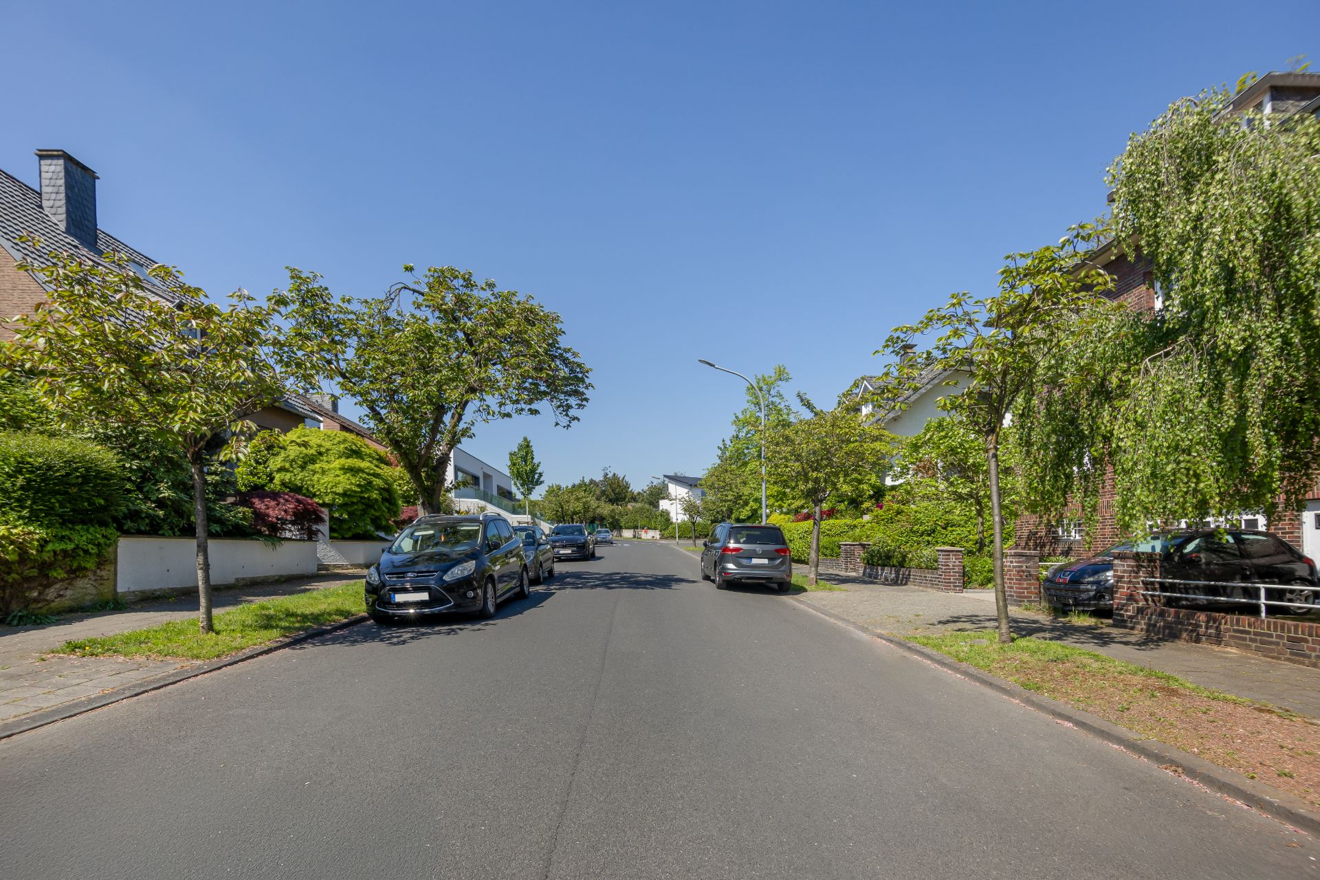 Ruhige Wohnstraße mit parkenden Autos, Bäumen und Häusern an einem sonnigen Tag.