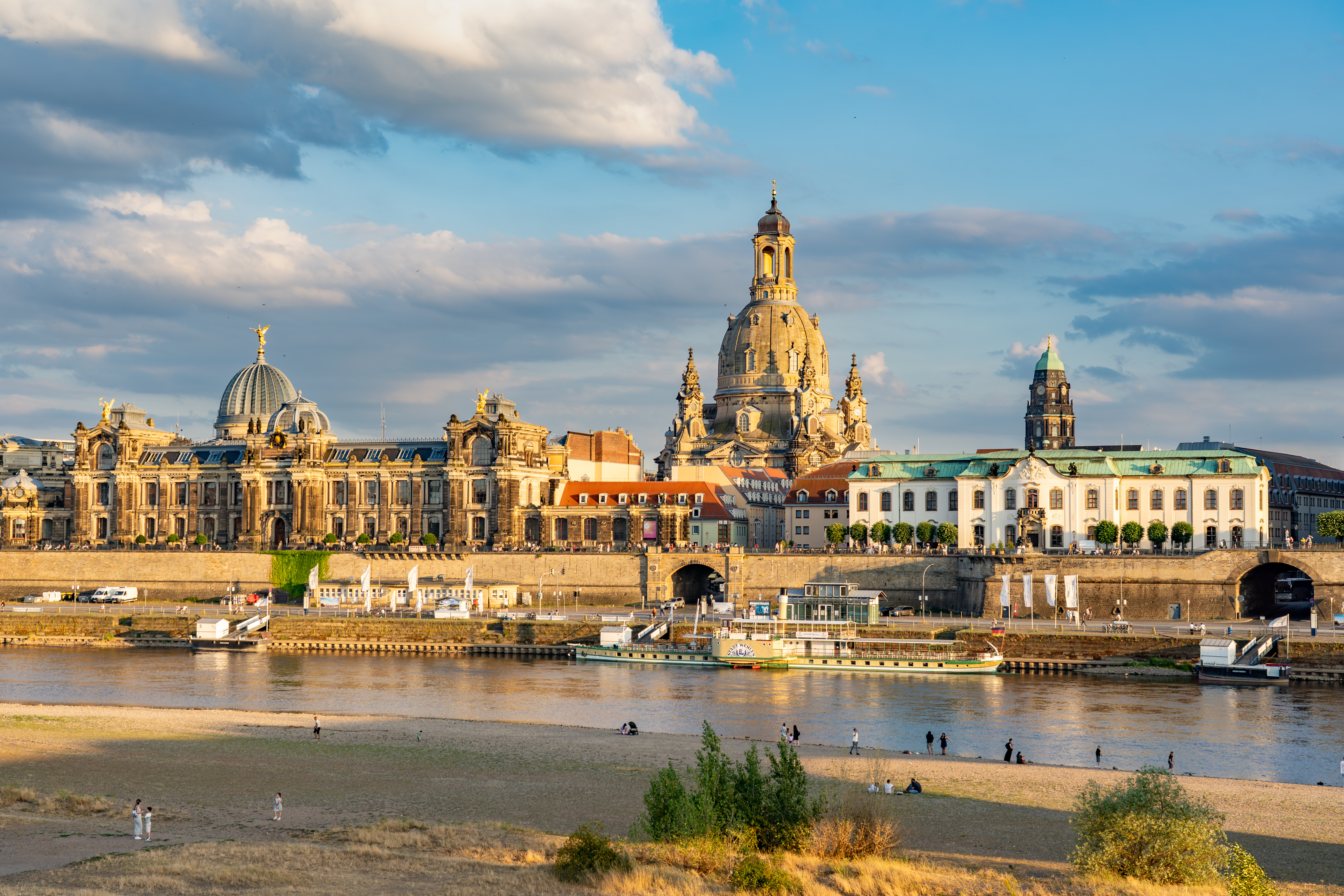 Stadtansicht mit historischen Gebäuden und Fluss im Vordergrund, Menschen am Ufer.