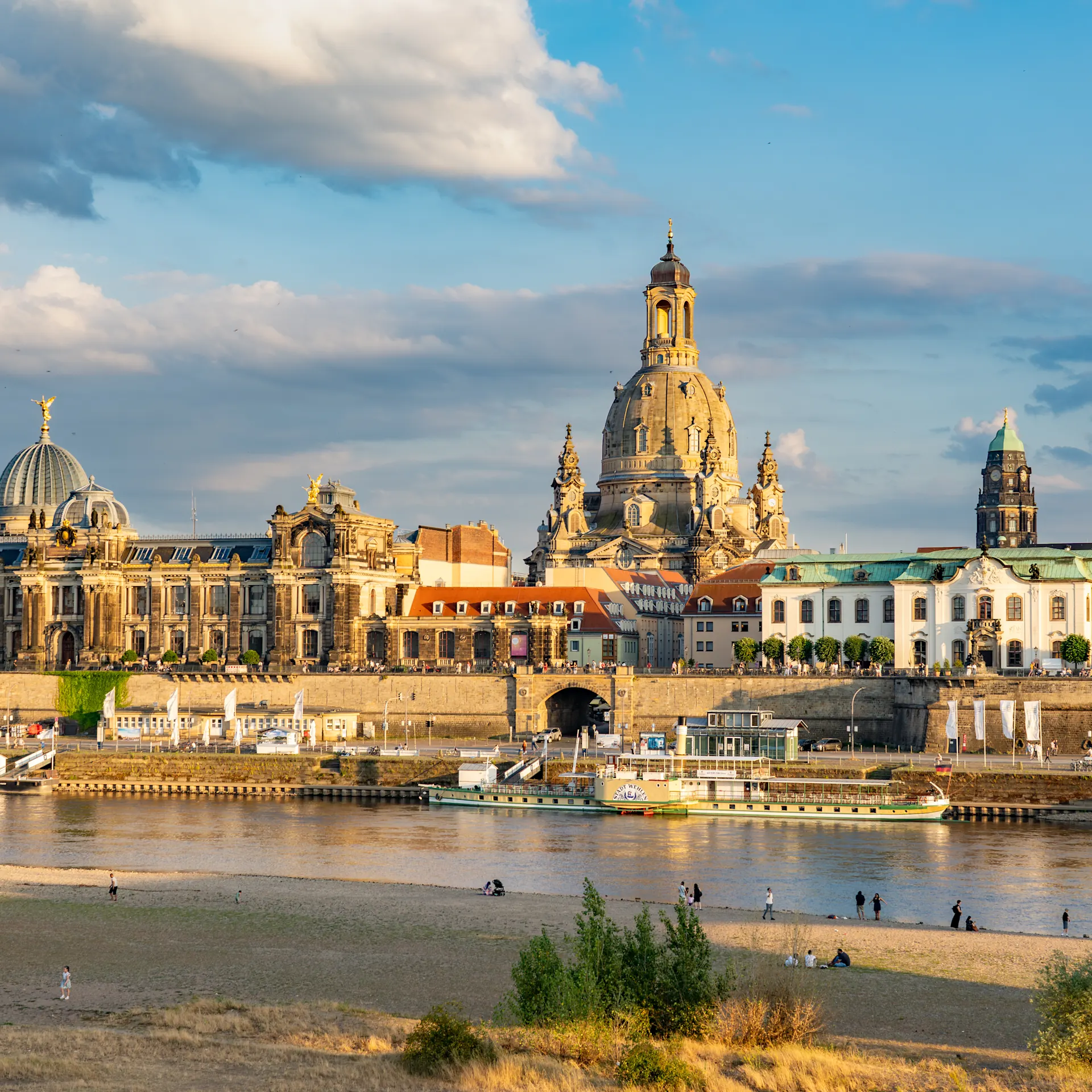 Stadtansicht mit historischen Gebäuden und Fluss im Vordergrund, Menschen am Ufer.