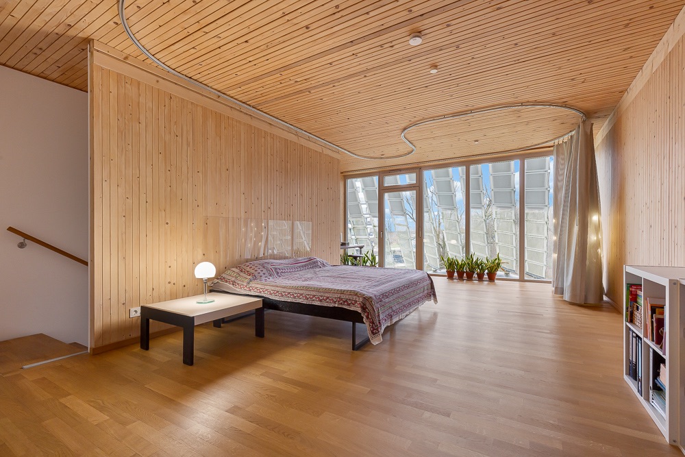 Helles Schlafzimmer mit Holzverkleidung, Doppelbett, Nachttischlampe und großen Fenstern mit Pflanzen.