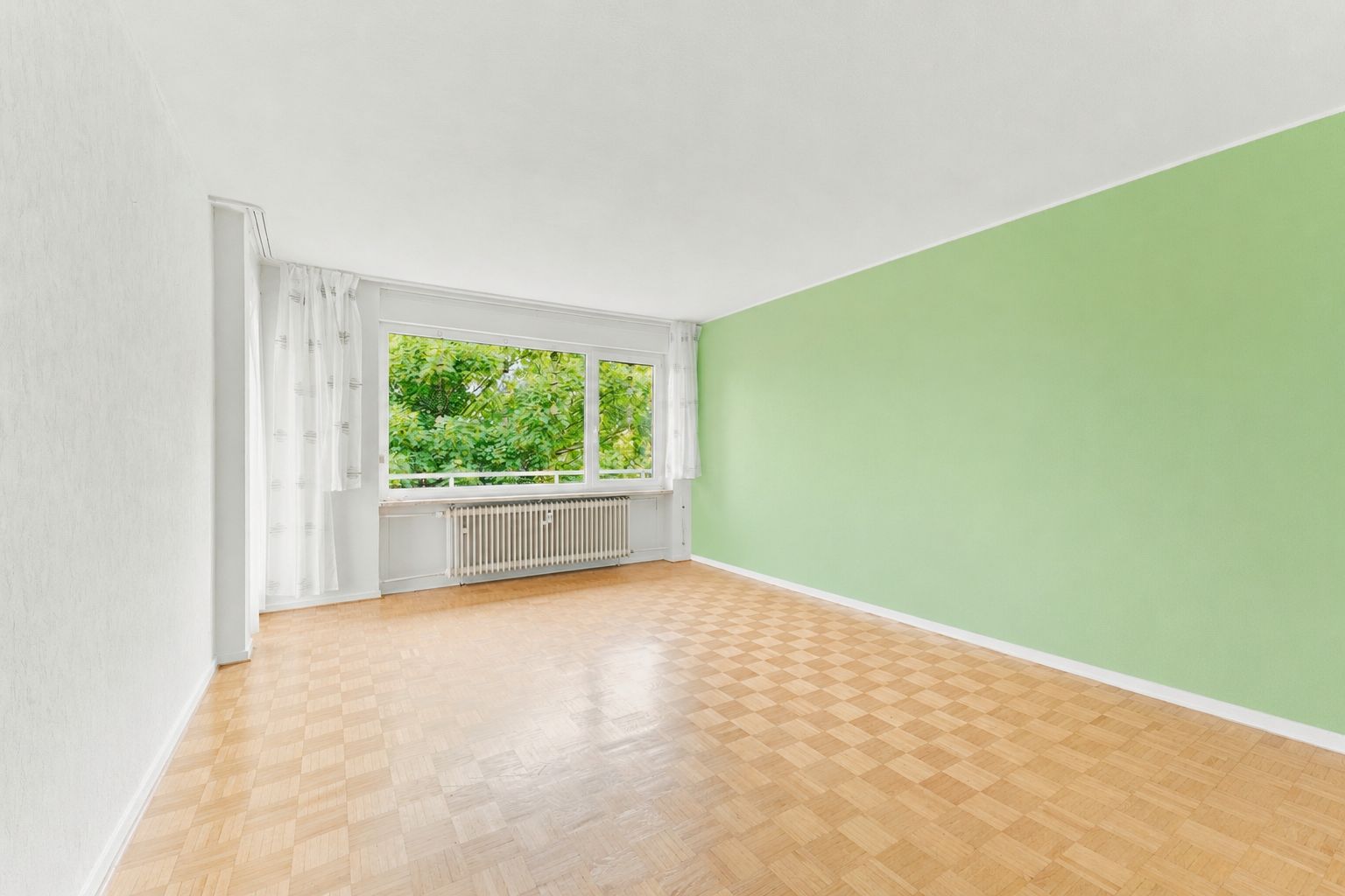 Leeres Wohnzimmer mit Parkettboden, grün gestrichener Wand und großem Fenster mit Blick ins Grüne.