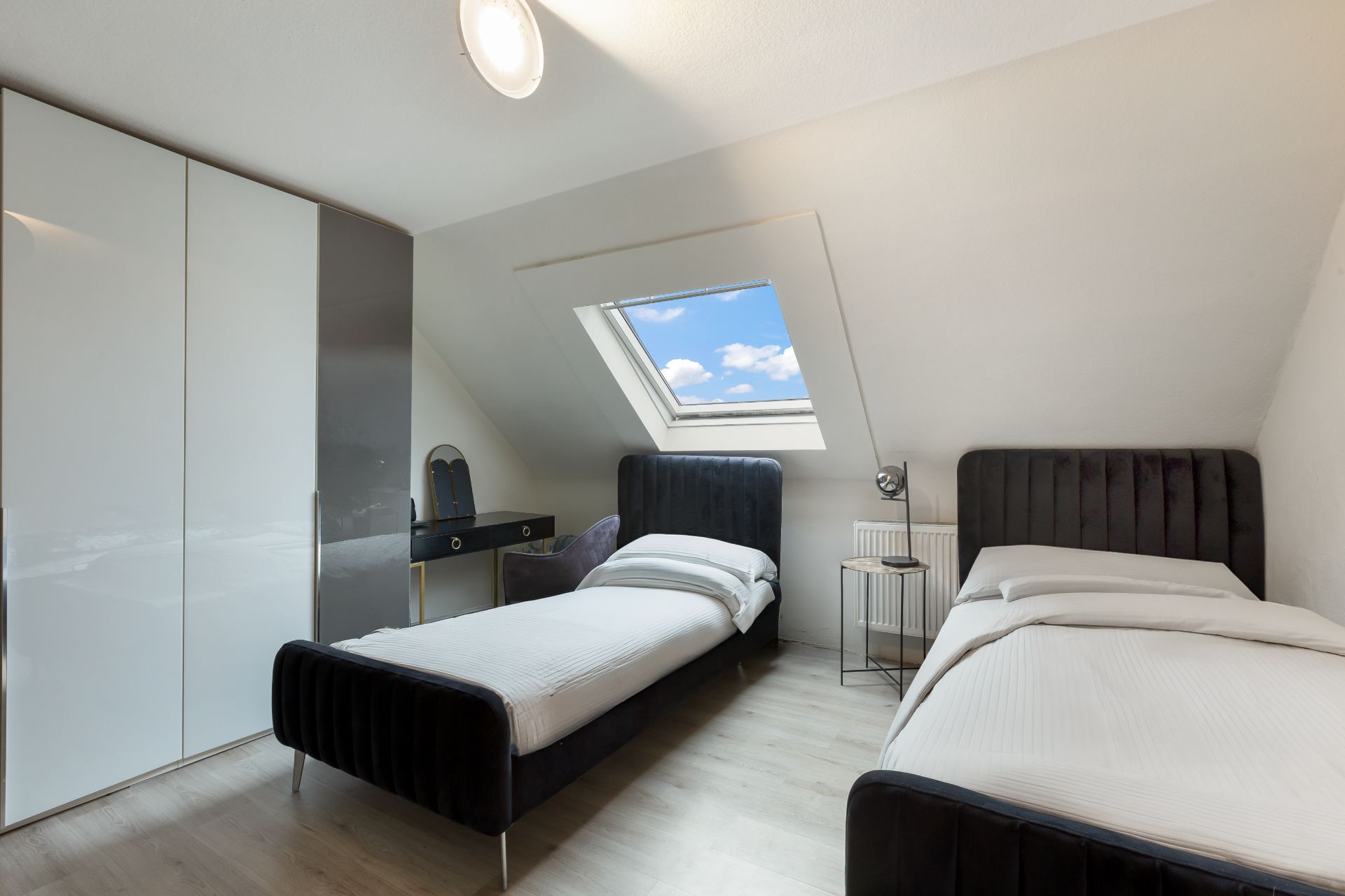 Helles Schlafzimmer mit zwei Einzelbetten, Kleiderschrank, Fenster und Schreibtisch unter Dachschräge.