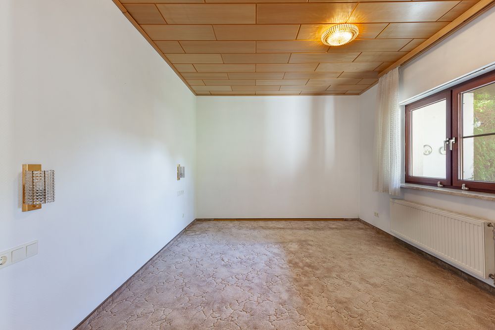 Leeres Zimmer mit Teppichboden, Holzdecke, großem Fenster und Heizkörper.