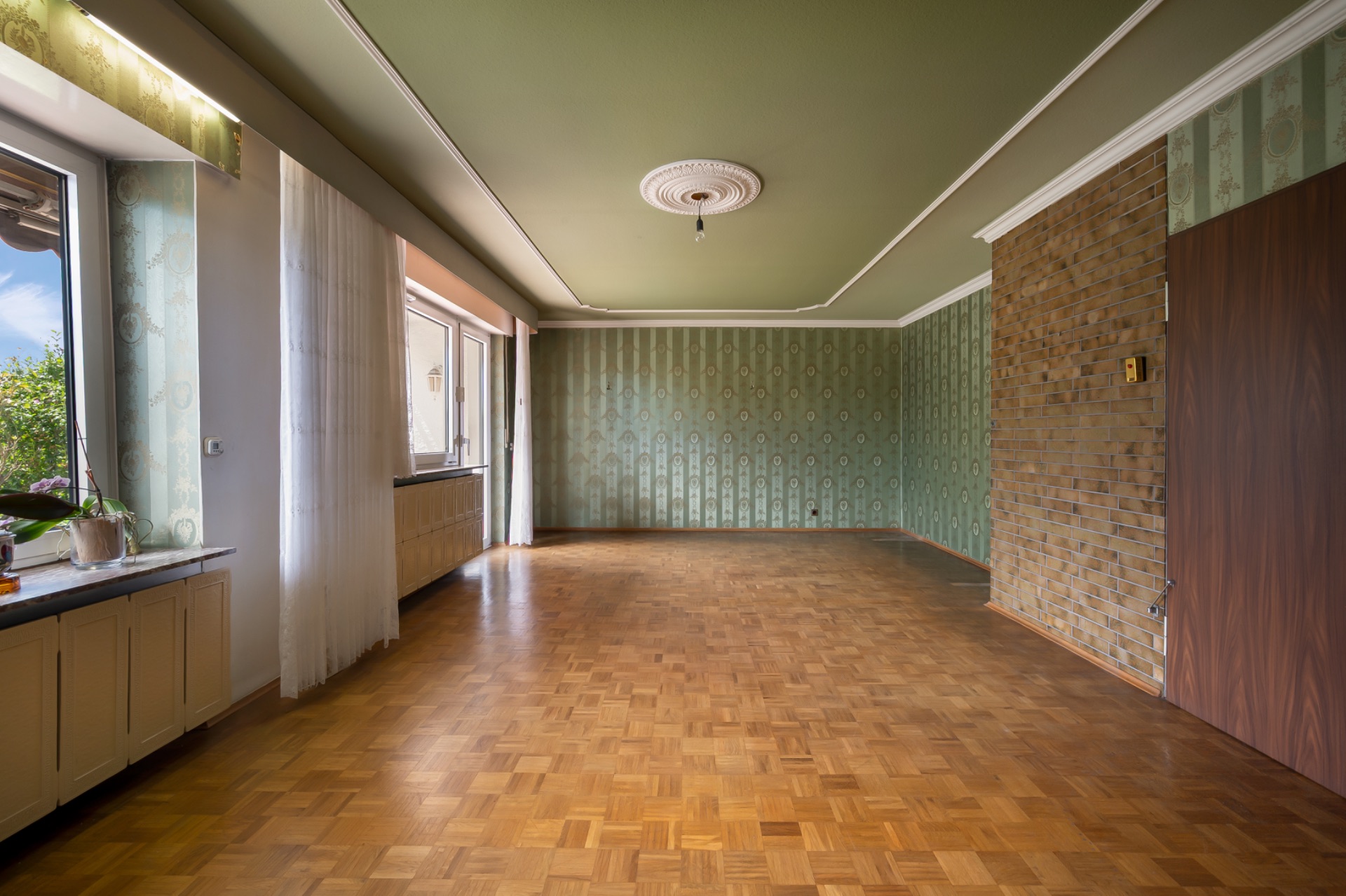 Leeres Wohnzimmer mit Parkettboden, großen Fenstern und grüner Tapete.