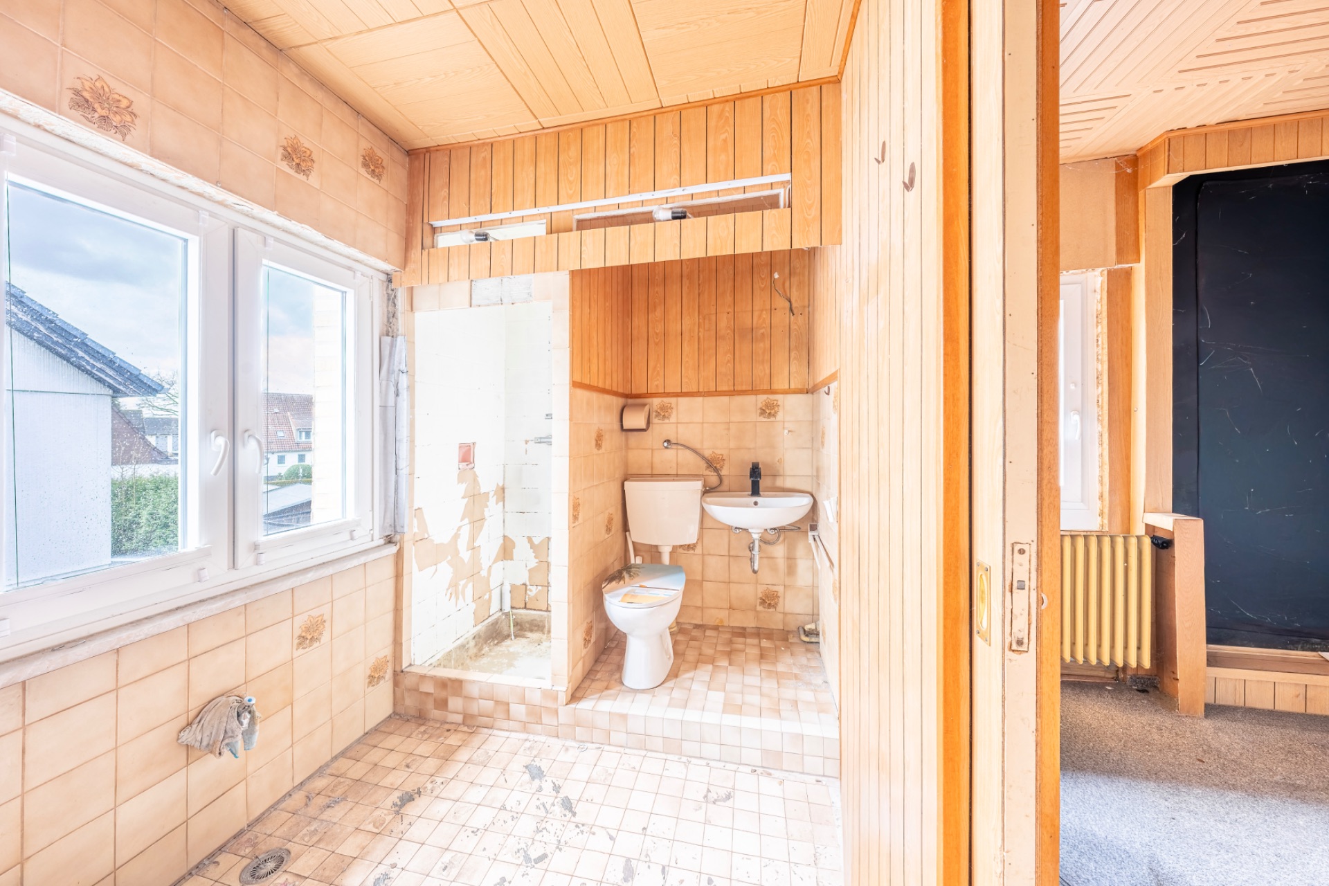 Kleines Badezimmer mit Toilette, Waschbecken und Dusche, Fliesen und Holzverkleidung, Fenster auf der linken Seite.
