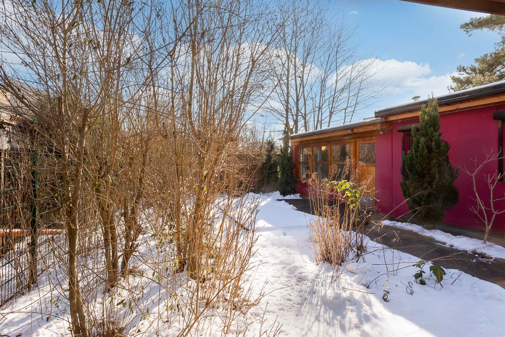 Garten mit schneebedecktem Boden neben einem roten Wohnhaus und kahlen Büschen.