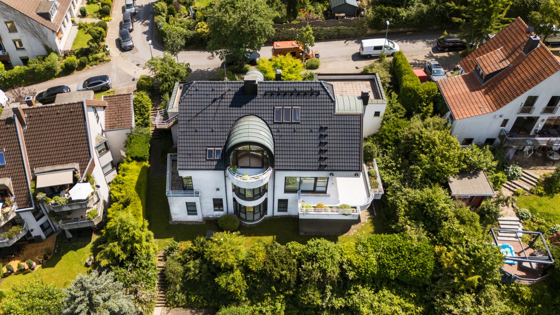 Luftaufnahme eines modernen Wohnhauses mit dunklem Dach und rundem Balkon, umgeben von Garten und Nachbarhäusern.