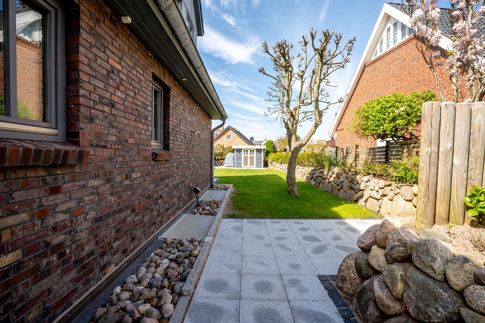 Gepflasterte Terrasse mit Steinen, angrenzendem Rasen und Baum neben einem Backsteinhaus.